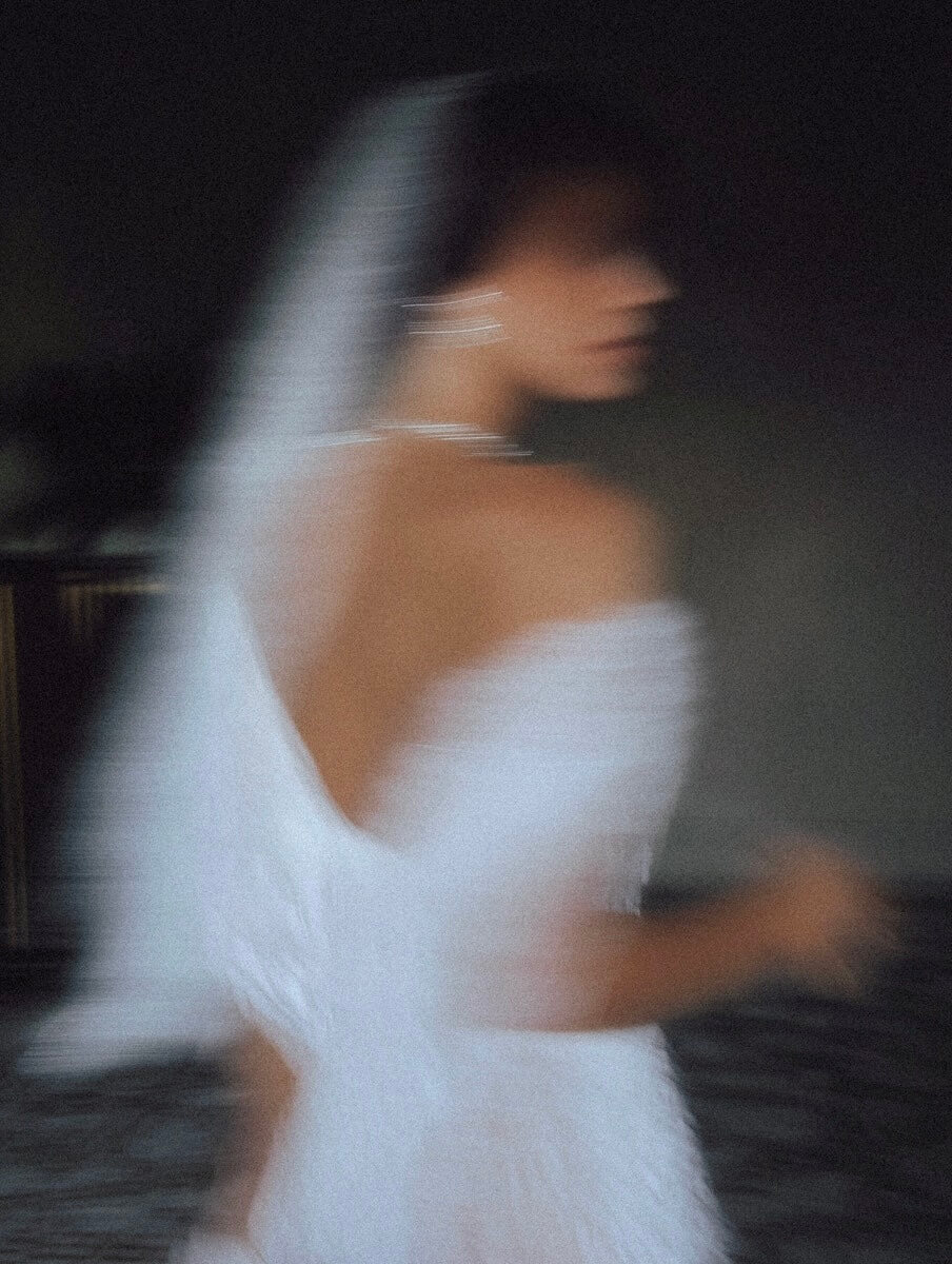 Artistic blurred photo of a bride in a white off-the-shoulder wedding dress with veil, turning away from the camera. Modern editorial bridal portrait.