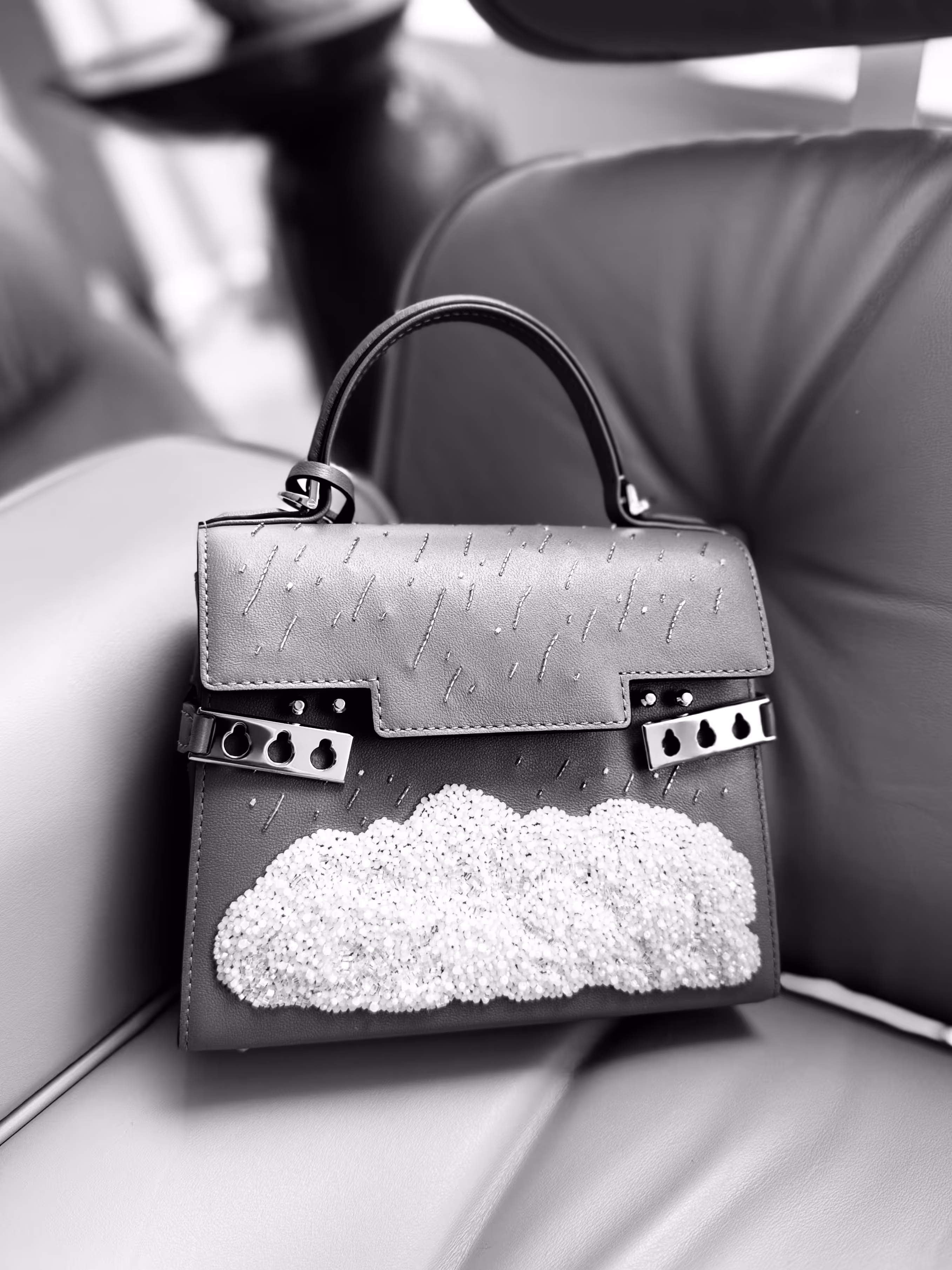 A black and white image of a handbag, Tempête Small Le Chant de l’Orage by Delvaux, resting on a leather seat.