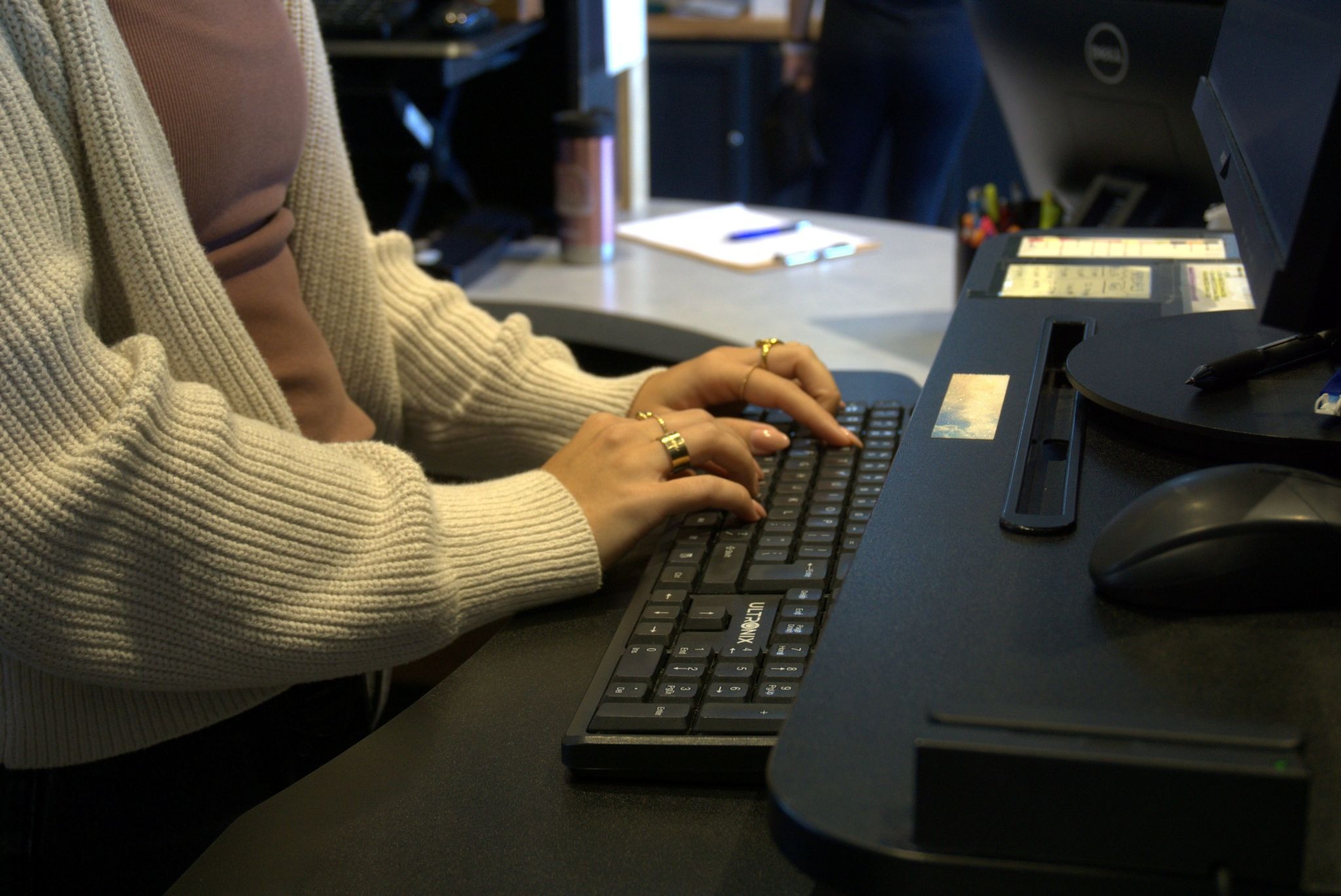 Person wearing a beige cardigan typing on a keyboard at a desktop computer in an office workspace, illustrating a typical seated posture while working at a desk.
