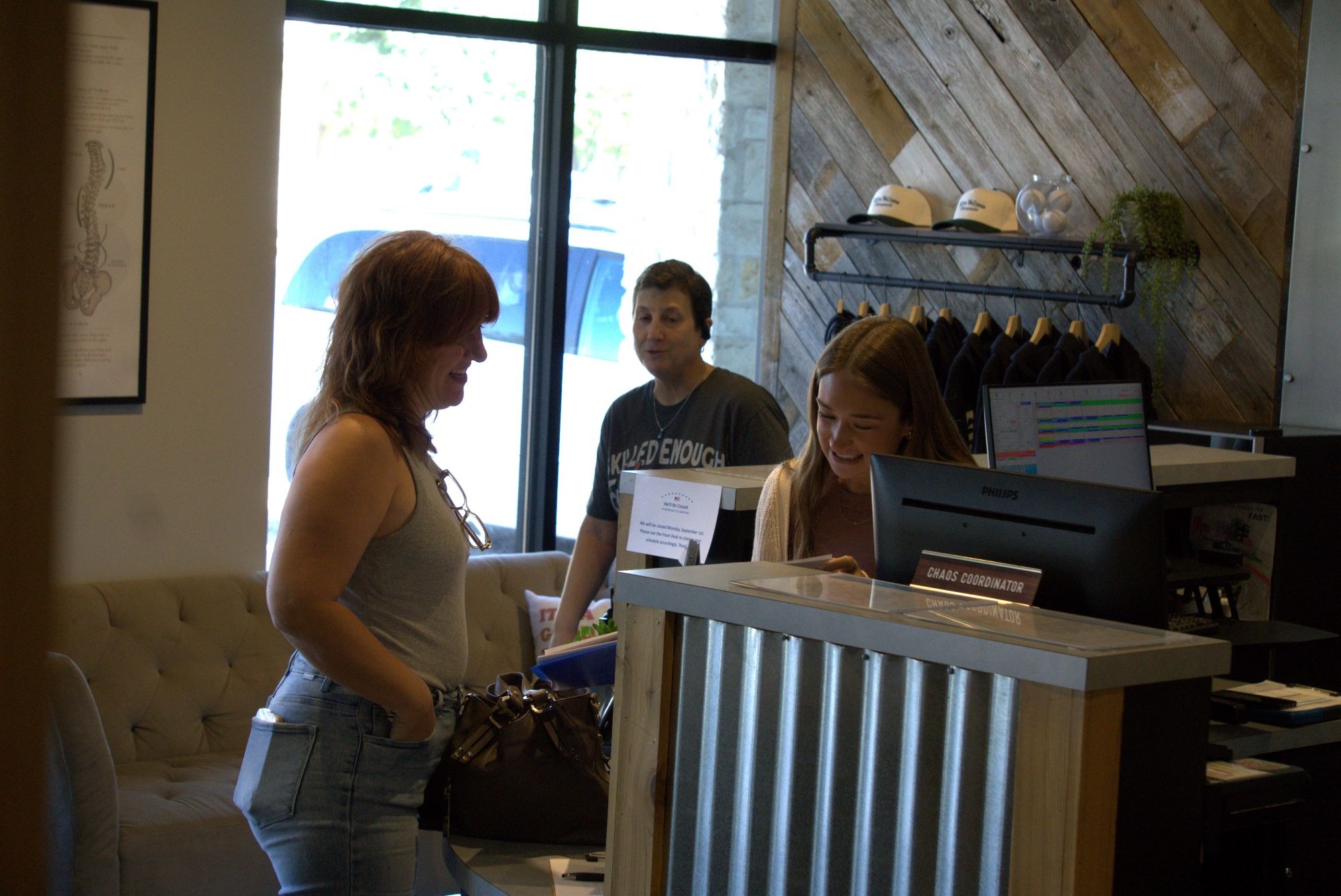 Patient checking in at the reception desk while speaking with a staff member, with another visitor waiting nearby in the bright and welcoming lobby of a chiropractic clinic.