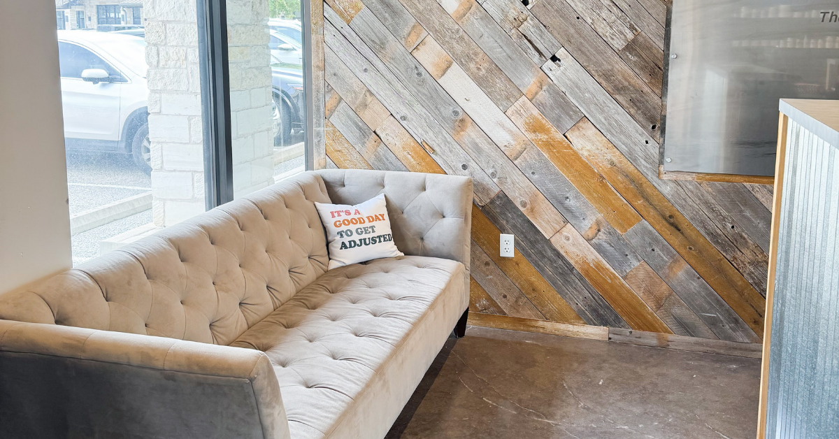 Comfortable waiting area at Elite Wellness Chiropractic featuring a tufted sofa, rustic wood accent wall, and natural light from large front windows.