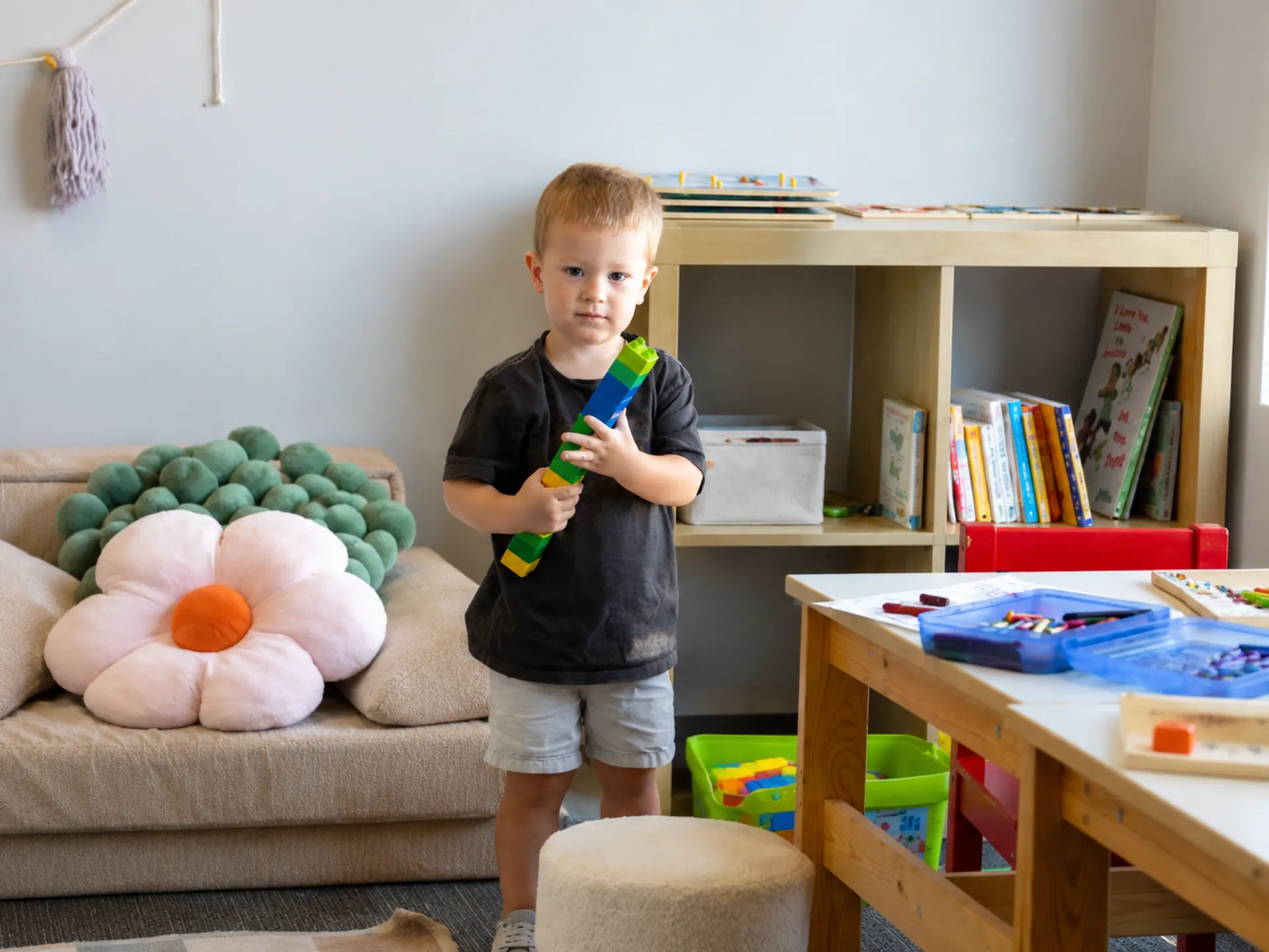 Toddler standing in a playroom holding a colorful toy with books, toys, and a table in the background.