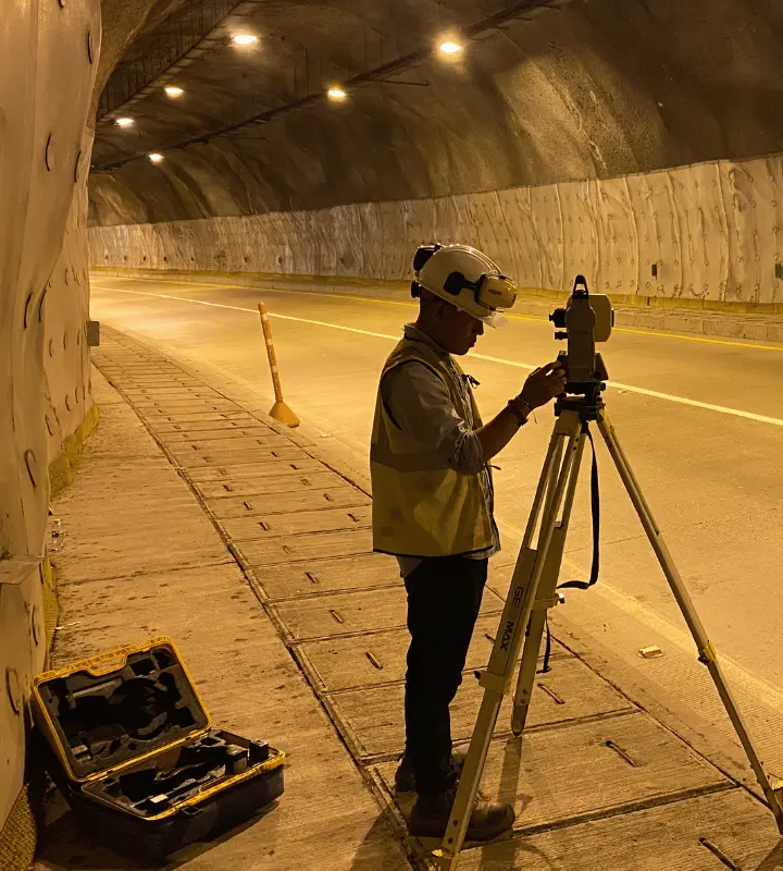 Trabajador con casco y chaleco realiza medición con equipo sobre trípode en túnel iluminado.