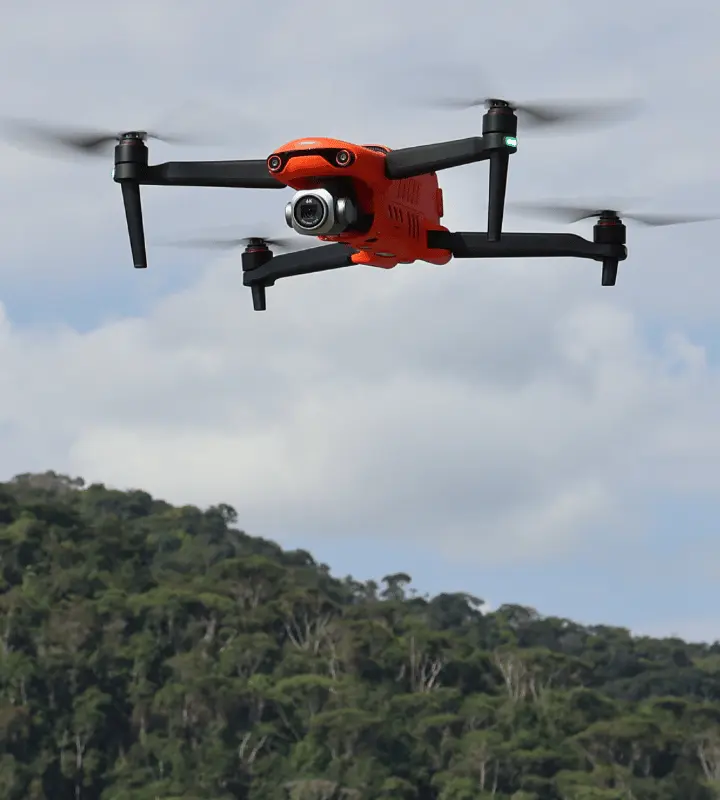 Dron naranja volando sobre árboles verdes con cielo nublado de fondo.