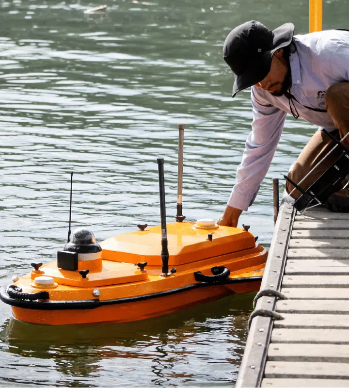 Hombre con sombrero negro ajusta un dron acuático naranja cerca de un muelle sobre agua.