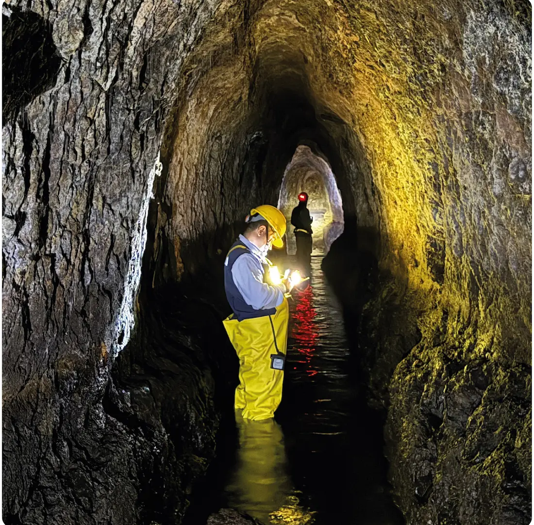 Dos personas con equipo de seguridad inspeccionan un túnel con agua hasta las rodillas.