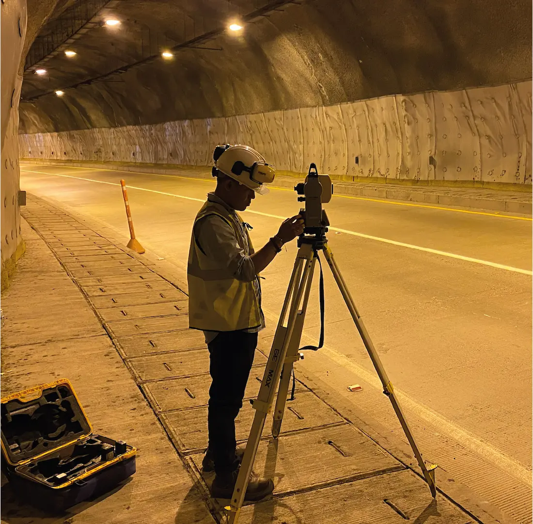 Trabajador con casco y chaleco usa un teodolito en un túnel iluminado durante una inspección.