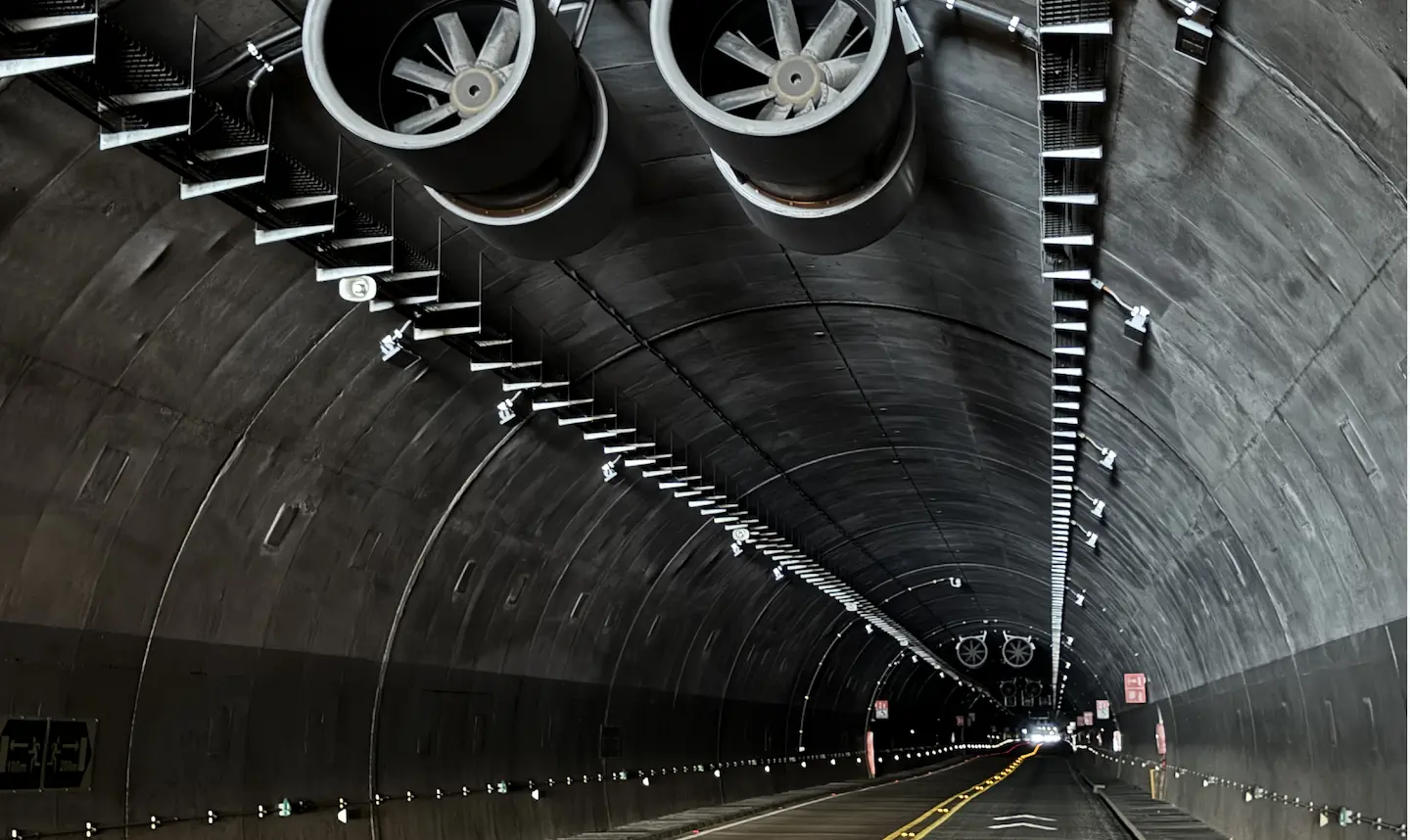 Interior de un túnel con ventiladores grandes y luces instaladas a lo largo de su techo.
