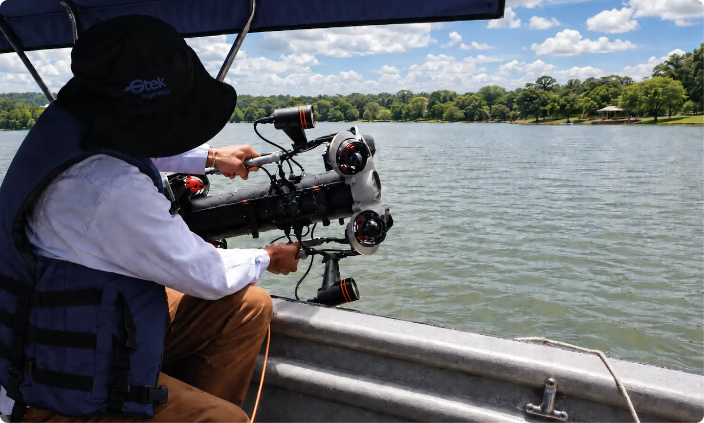 Persona en bote sostiene equipo de inspección submarina sobre un lago con árboles y cielo nublado.