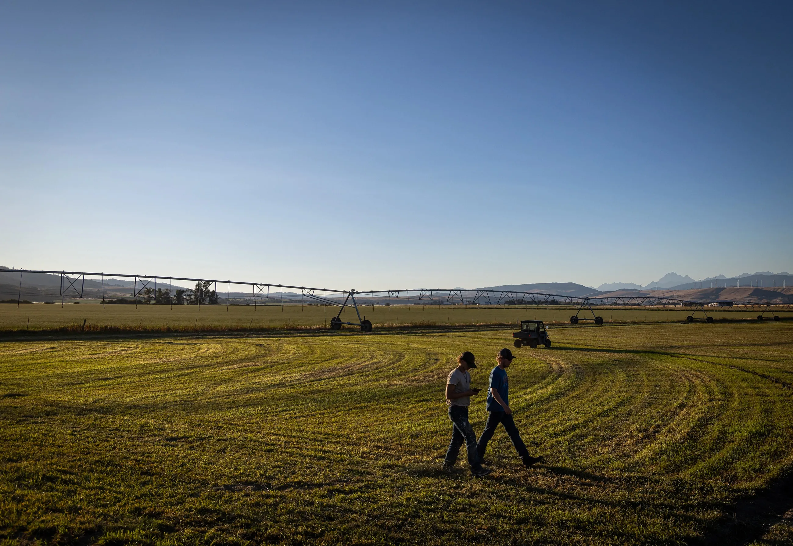 Two men walk across a large green field with farming irrigation equipment and mountains in the background under a clear blue sky.