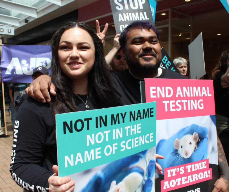 Two people at a protest holding signs that say 'Not in my name, not in the name of science' and 'End animal testing, It's time Aotearoa!' with an image of a white lab mouse.
