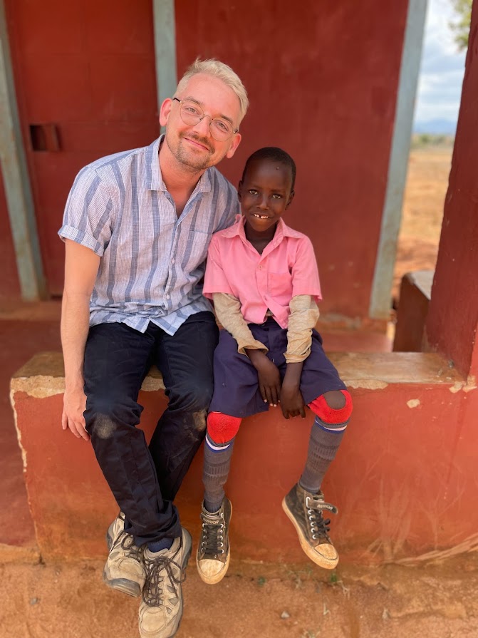Bonar Bell sitting next to a smiling child in a pink shirt and knee pads on a red concrete bench in Zambia.