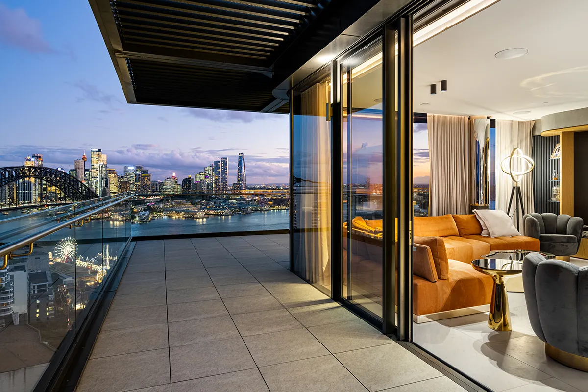 A modern luxury apartment in Sydney with an open living room and large balcony overlooks Sydney skyline and tthe Harbour Bridge at sunset. Glass walls provide panoramic views, and stylish furniture decorates the interior.