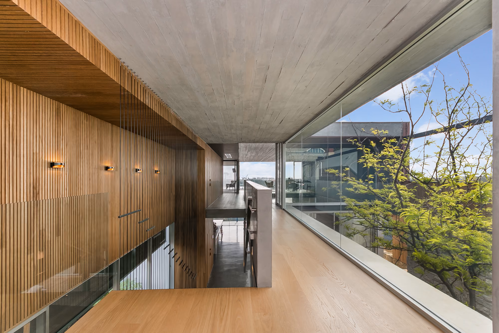 Modern interior hallway with wooden slat walls, concrete ceiling, light wood floor, and large glass window overlooking green tree and exterior architecture.