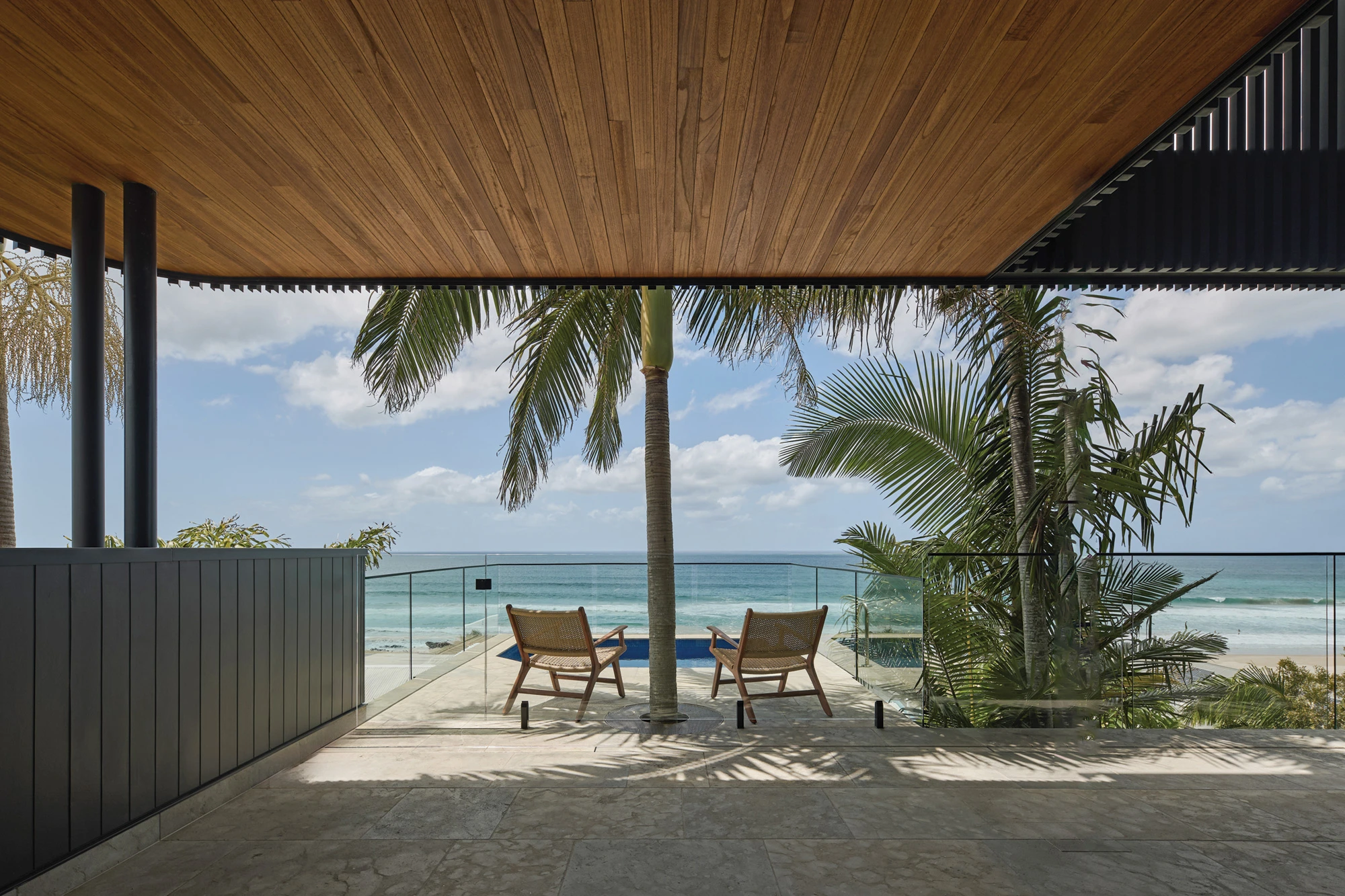 Two woven lounge chairs facing the ocean on a patio with glass railing and palm trees.