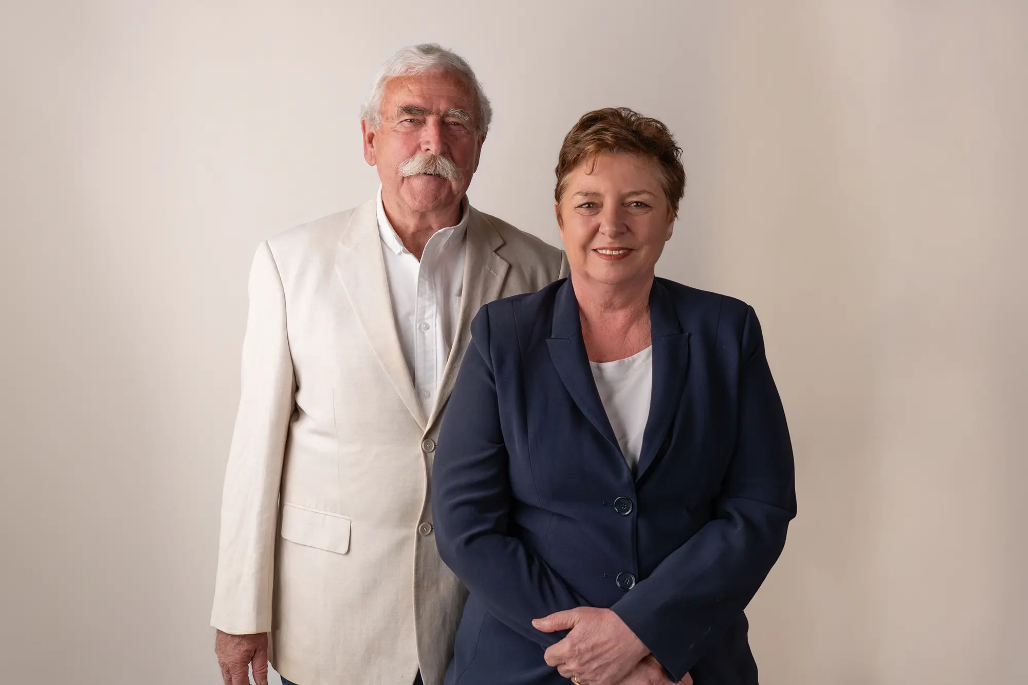 Smiling mature couple standing side by side against a beige background; man with white hair and mustache in a light beige blazer, woman with short hair in a navy blazer.