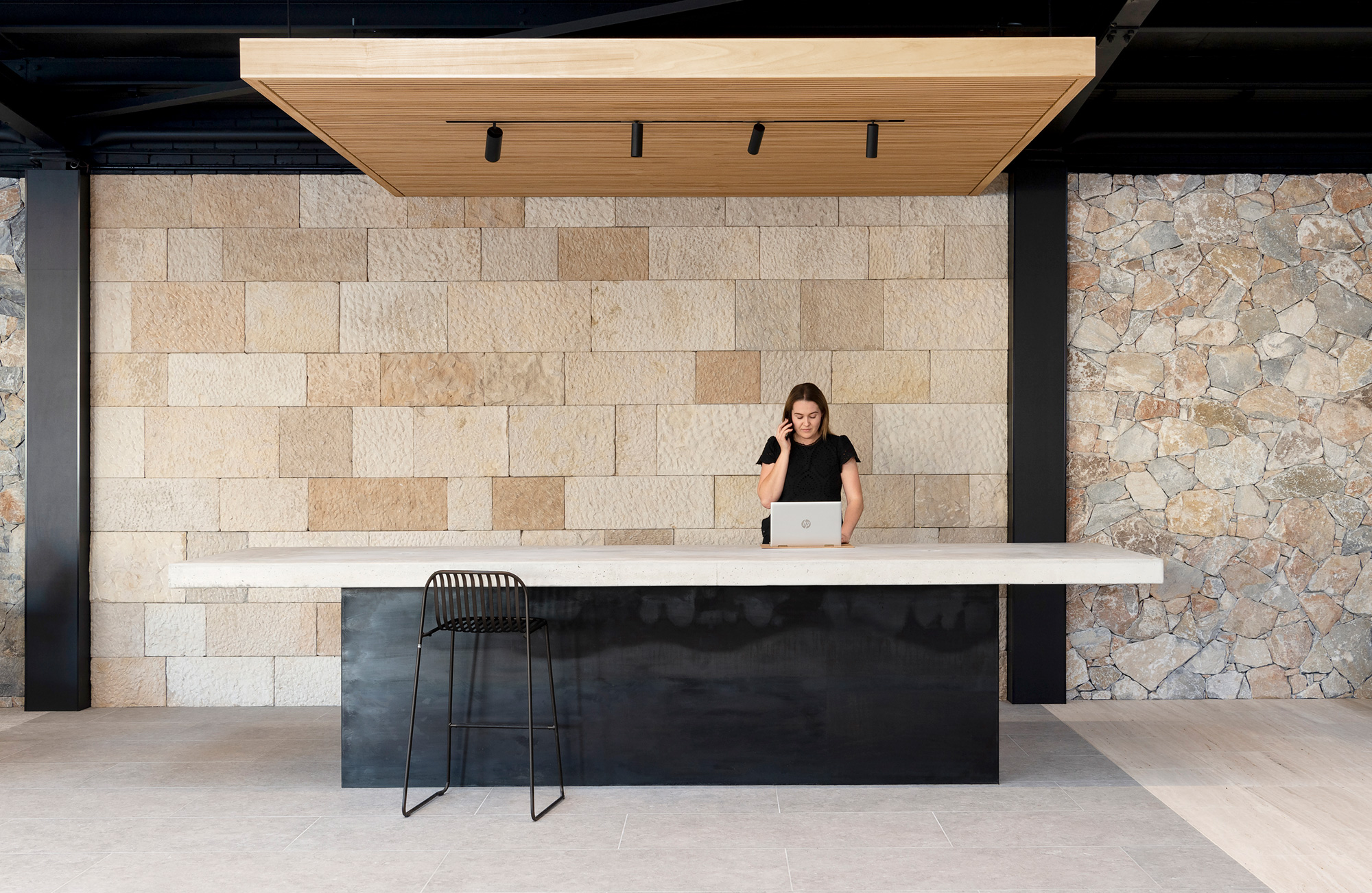 Woman talking on phone and working on laptop at a large black and white reception desk with stone walls in the background.