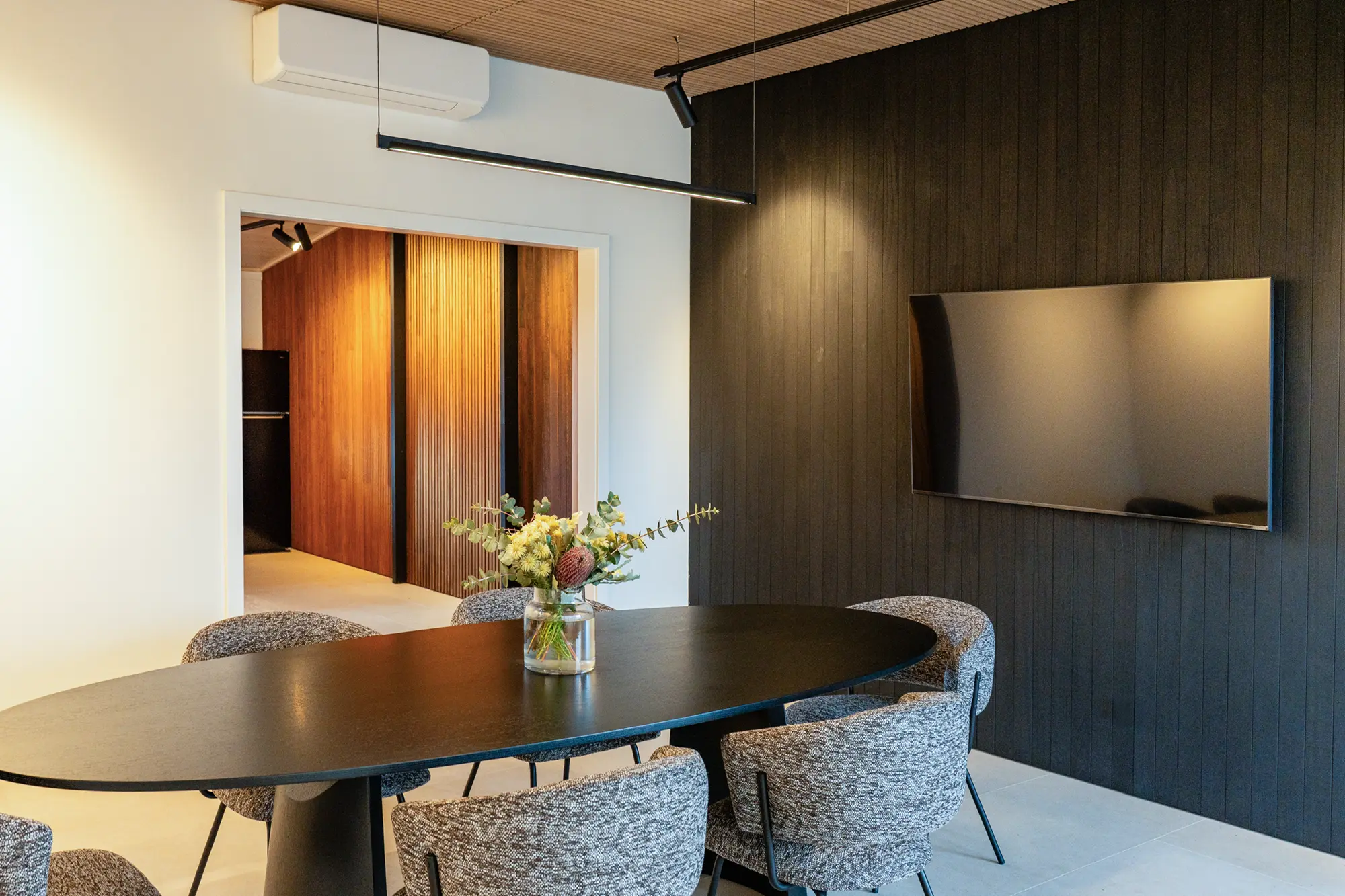 Modern dining room with an oval black table, six gray upholstered chairs, a vase with flowers in the center, a wall-mounted TV, and wood-paneled accent walls.