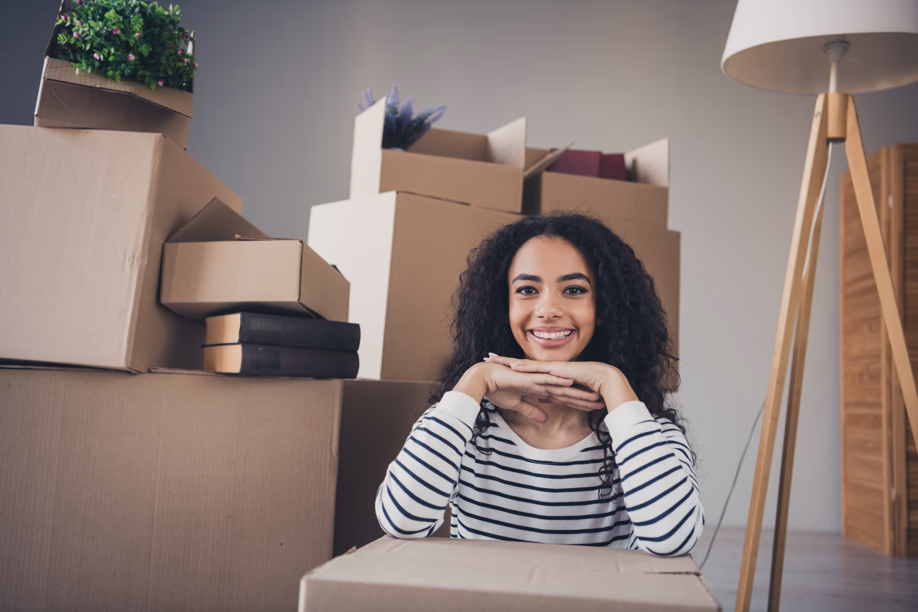 A young woman with curly hair smiling brightly while sitting among stacks of moving boxes, representing the excitement of moving into her new home at The Rook in Orlando, FL.