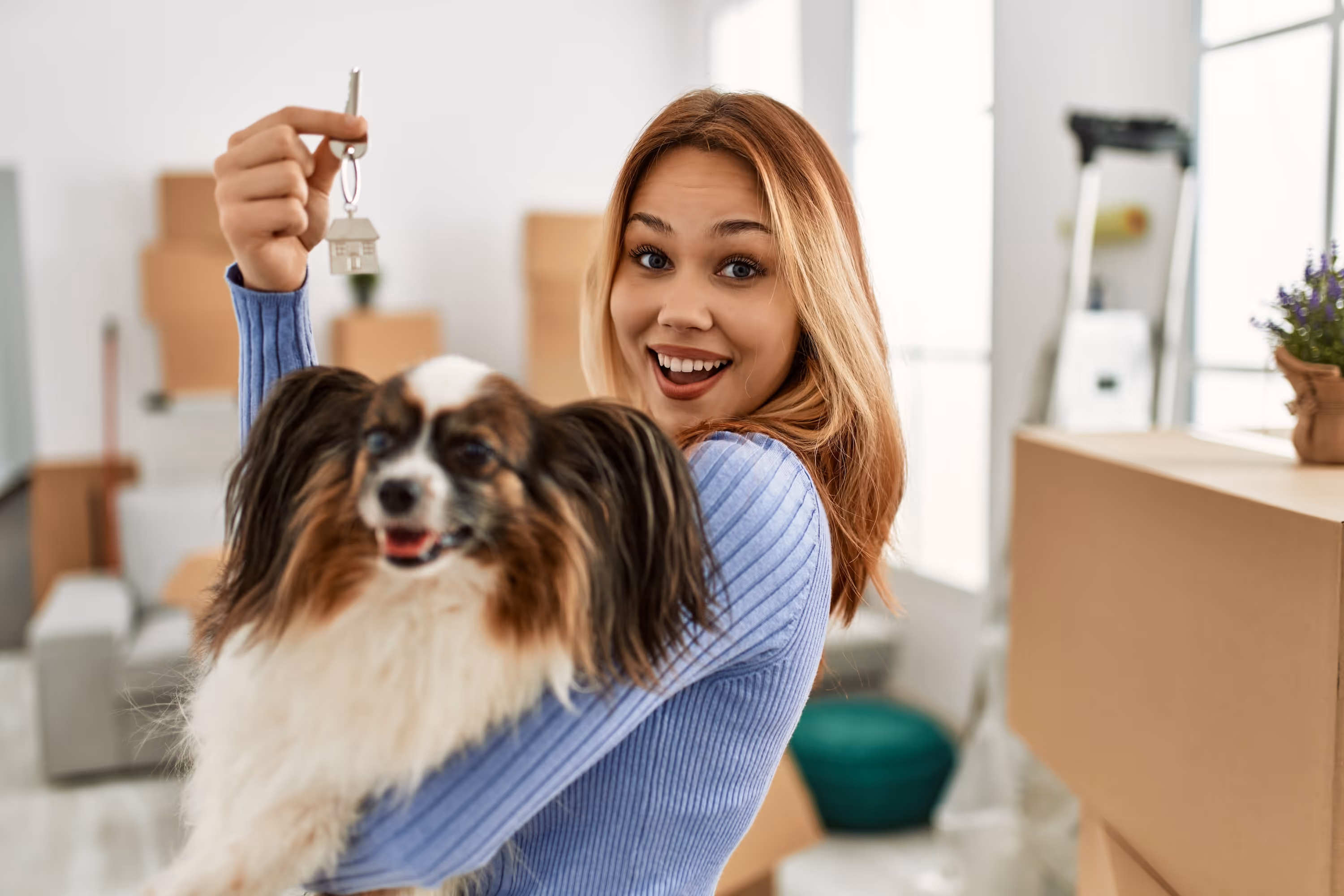 Happy woman holding up new apartment keys and hugging a small dog in a room at The Rook with moving boxes.