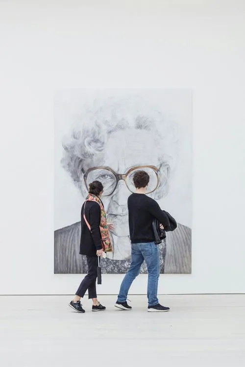 Two people observing a large portrait of an elderly woman in an art gallery