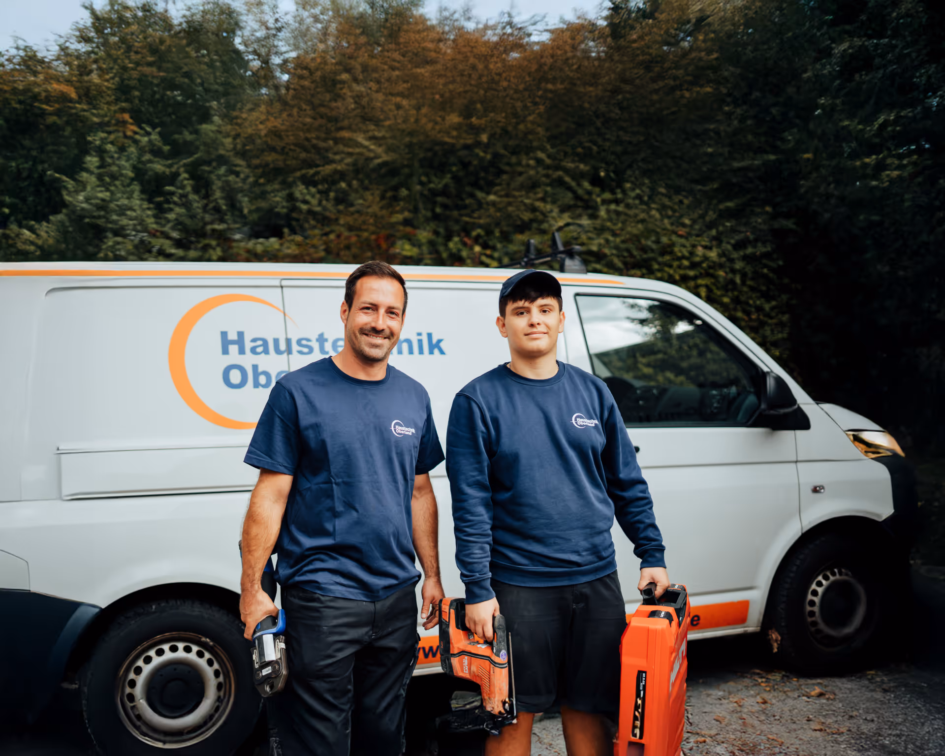 Zwei HTO Handwerker mit Werkzeugkoffern und Firmenwagen im Hintergrund