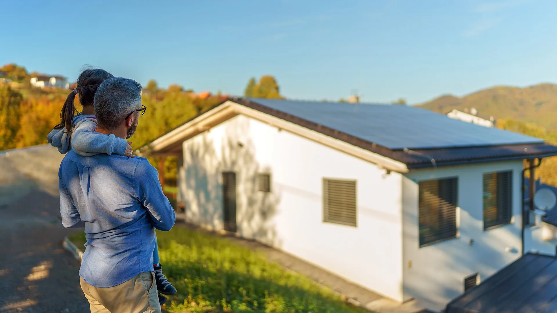 Eine Person hält ein Kind im Arm und blickt an einem sonnigen Tag auf ein Haus mit Sonnenkollektoren, umgeben von Grün und Herbstfarben.