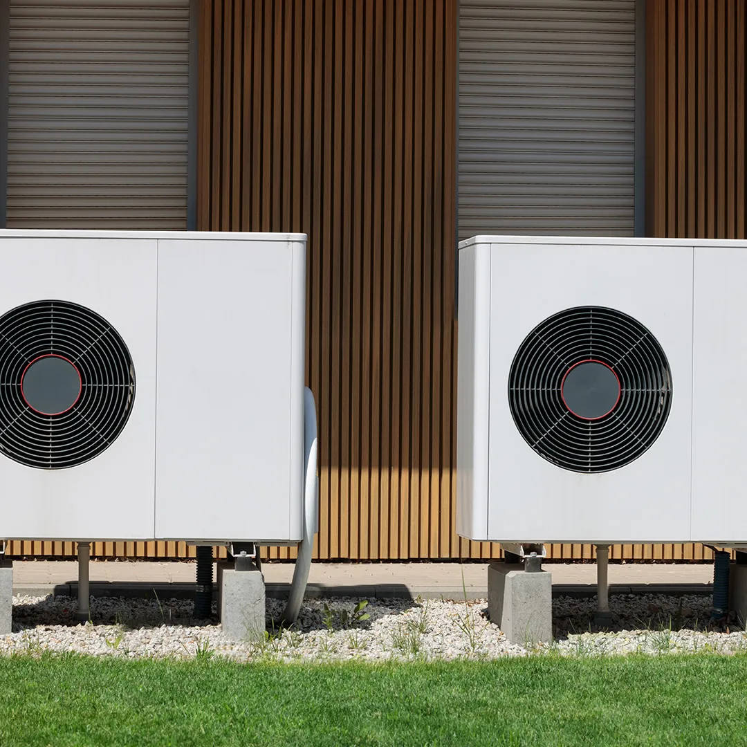 Zwei weiße Wärmepumpen auf Betonsockeln, umgeben von Kies und Gras, vor einer Holzwand.