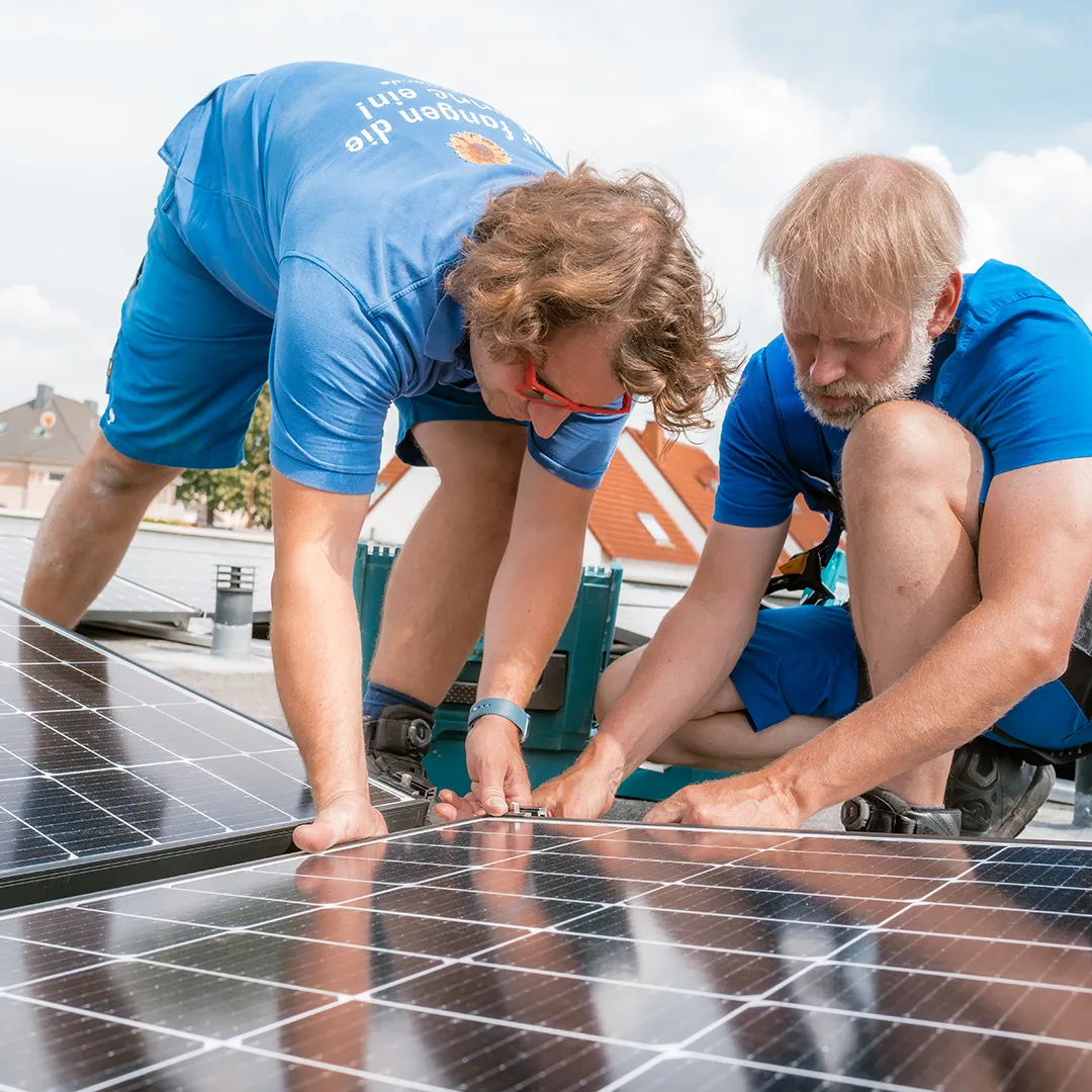 Zwei Arbeiter in blauer Kleidung installieren Sonnenkollektoren auf einem Dach und konzentrieren sich auf die Befestigung der Komponenten. Im Hintergrund sind der klare Himmel und Häuser zu sehen.
