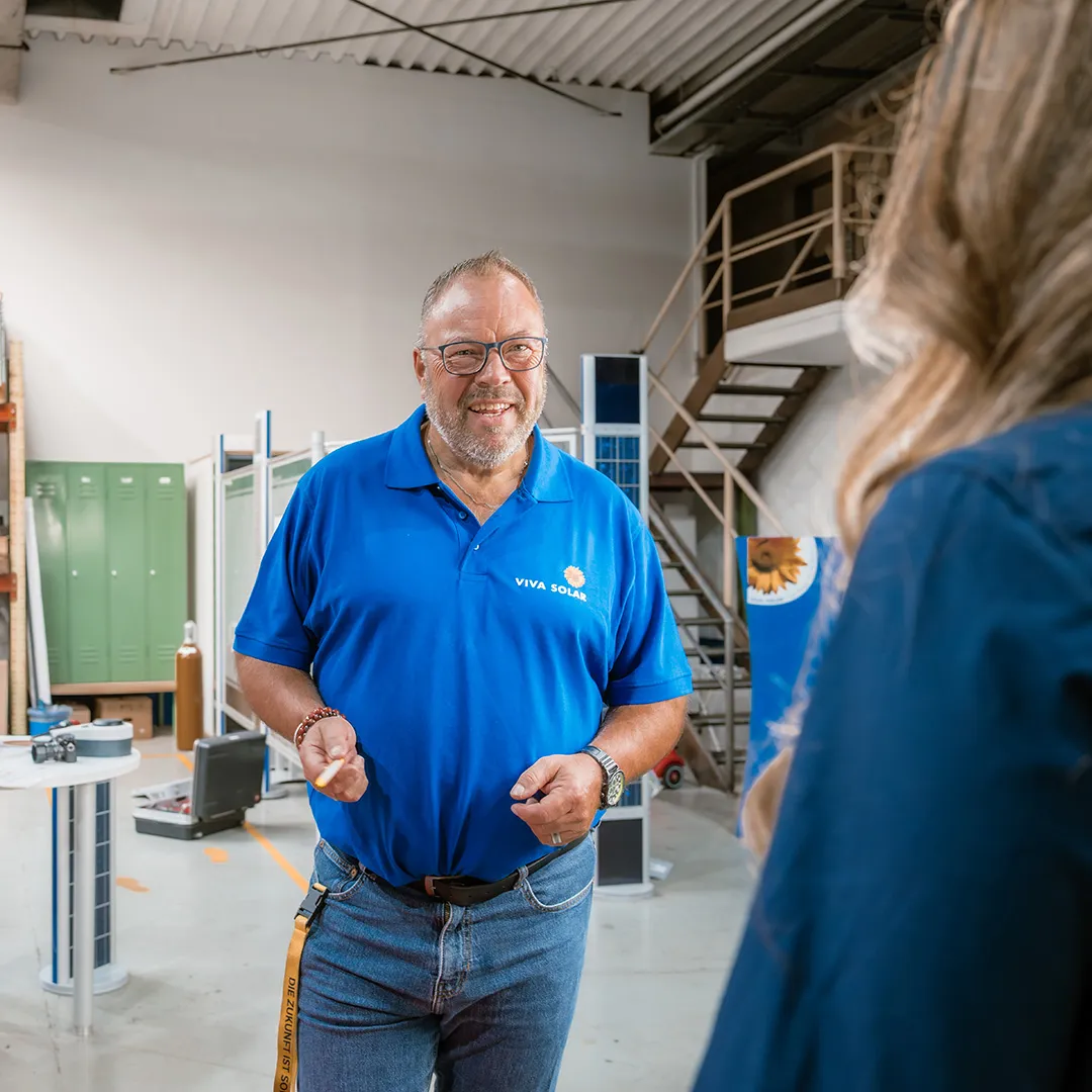 Ein lächelnder Mann in einem blauen VIVA SOLAR-Poloshirt steht in einem Industriegebäude und unterhält sich mit einer Person außerhalb des Bildausschnitts.