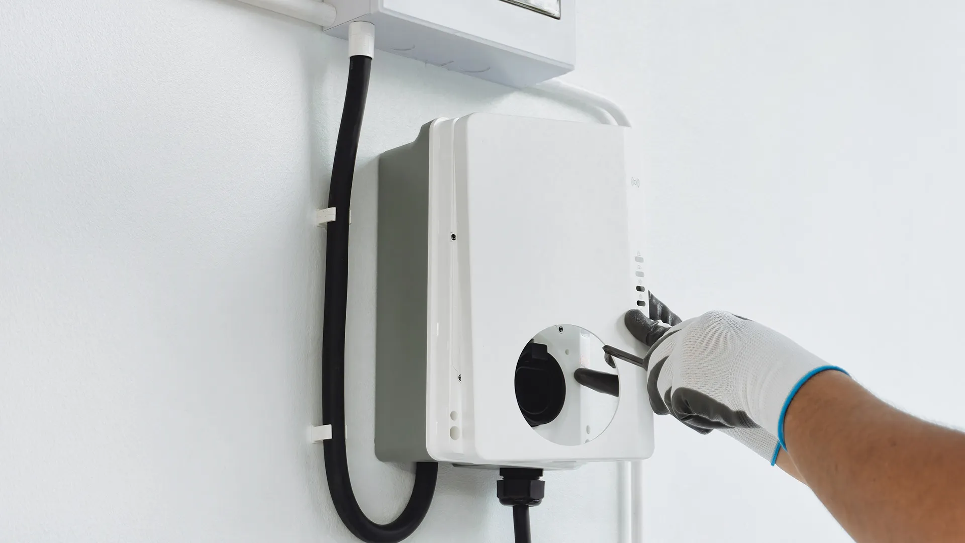 Eine Hand in einem Handschuh bedient ein weißes elektronisches Gerät, das an einer Wand montiert und über Kabel angeschlossen ist.