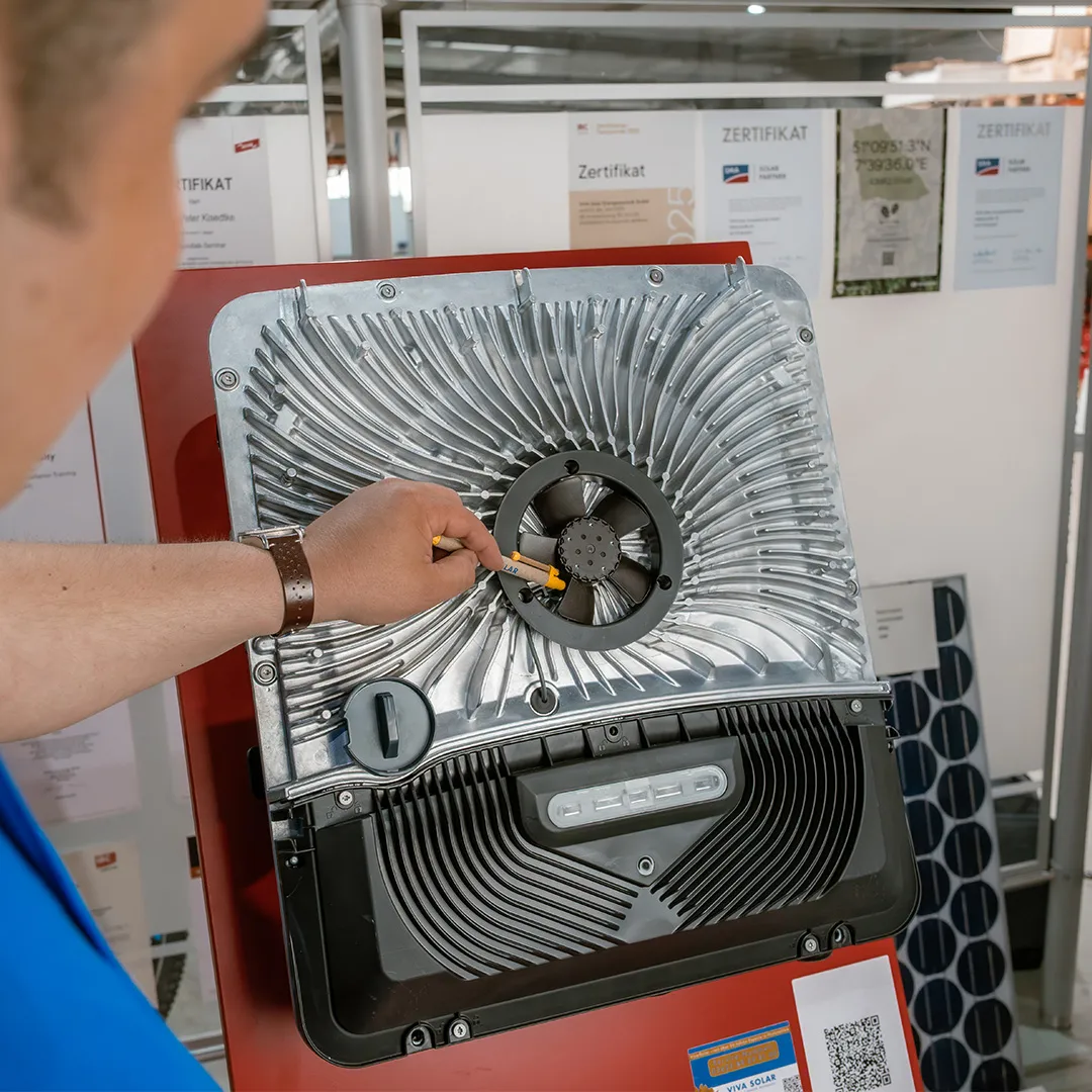 Eine Person zeigt mit einem Bleistift auf ein metallisches Bauteil eines Solarpanel-Displays, im Hintergrund sind Informationsplakate zu sehen.