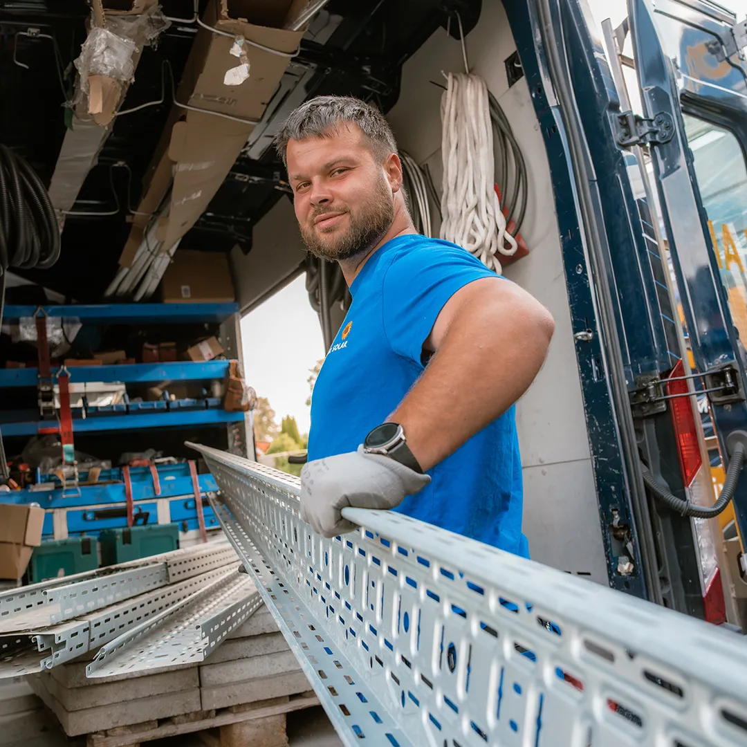 Ein Arbeiter in einem blauen Hemd hält ein langes Metallstück in der Hand, umgeben von Werkzeugen und Geräten in einem Lieferwagen. Natürliches Licht unterstreicht die Szene.
