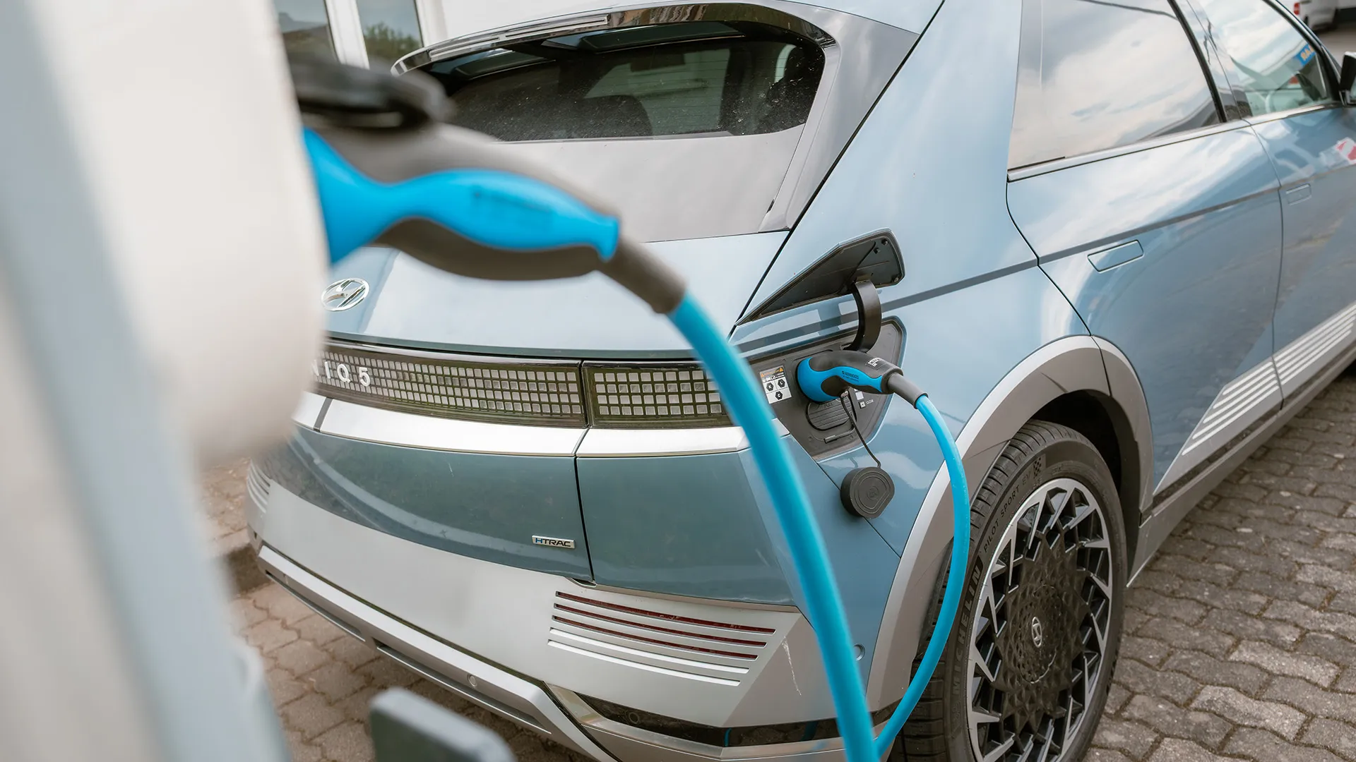 Ein blaues Elektroauto wird an einer Ladestation aufgeladen, wobei ein hellblaues Ladekabel an seinem Heck angeschlossen ist.
