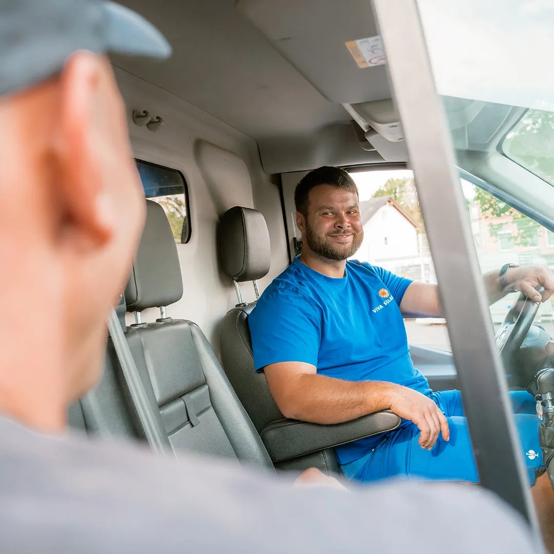 Ein lächelnder Mann in einem blauen Hemd sitzt auf dem Fahrersitz eines Kleinbusses und unterhält sich mit einer anderen Person außerhalb des Fahrzeugs.
