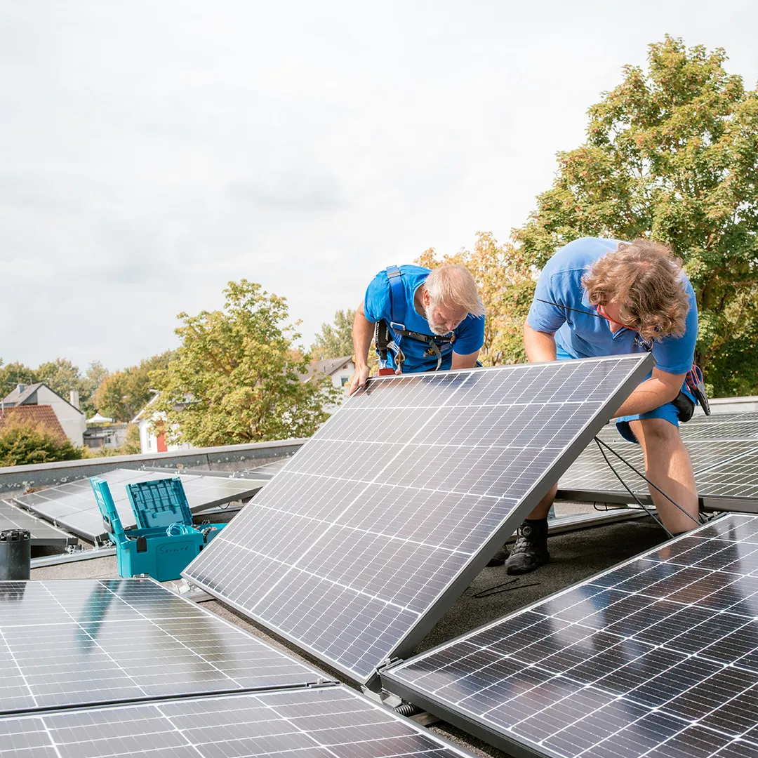 Zwei Arbeiter installieren Sonnenkollektoren auf einem Dach, im Hintergrund sind grüne Bäume und Häuser zu sehen. In der Nähe liegen Werkzeuge herum.