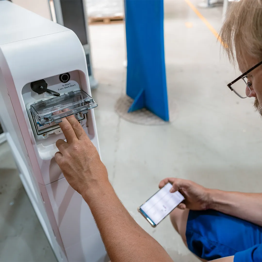 A person interacts with a machine, using one hand to press a button while holding a smartphone in the other, in an industrial setting.