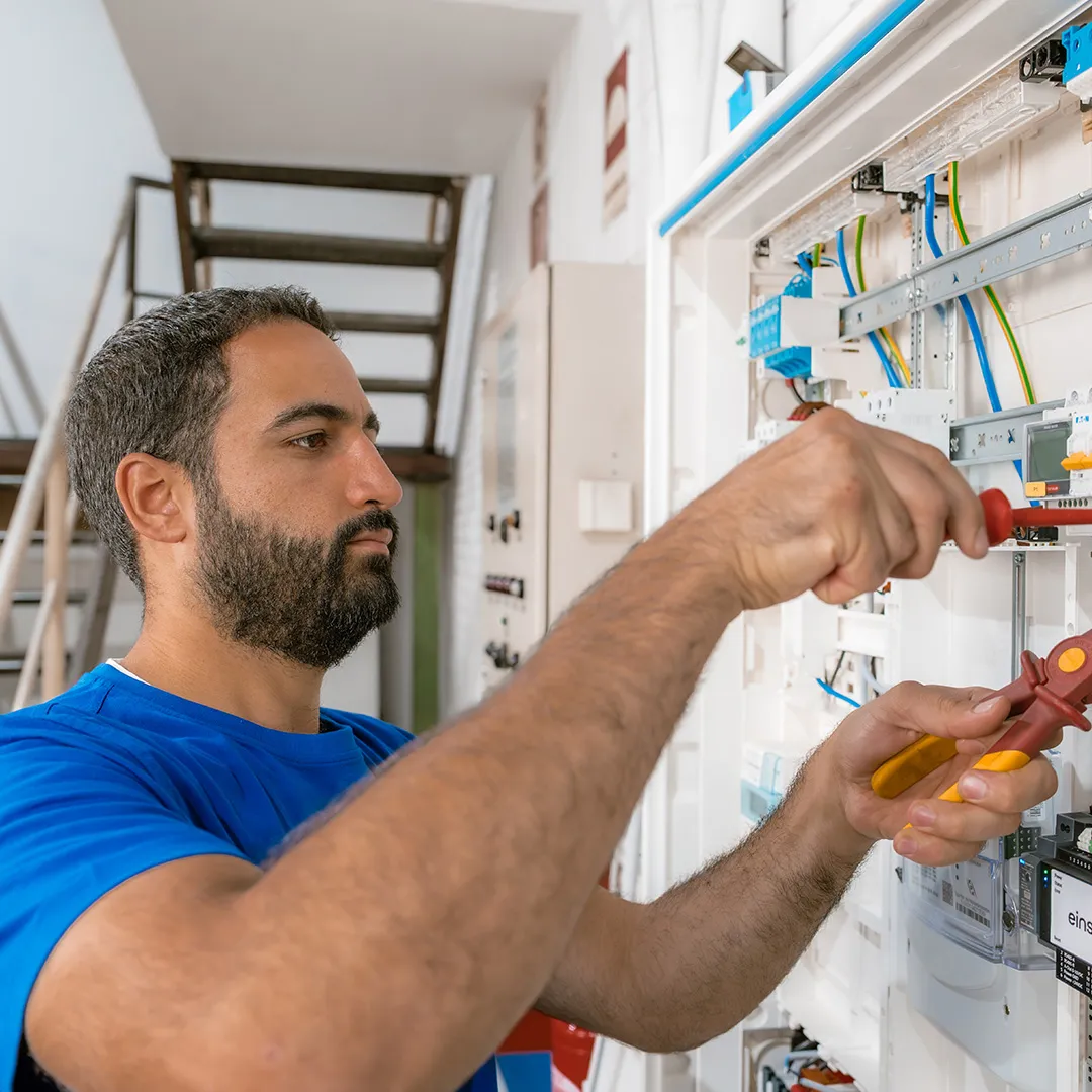 Ein Mann in einem blauen Hemd arbeitet mit Werkzeugen an elektrischen Geräten und konzentriert sich dabei auf ein Bedienfeld in einer hellen Innenraumumgebung.