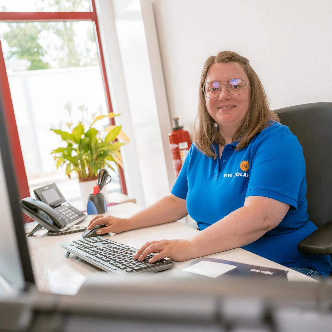 Eine Frau in einem blauen Polohemd sitzt an einem Schreibtisch mit einem Computer, einer Tastatur und Pflanzen in einem hellen Büroraum.