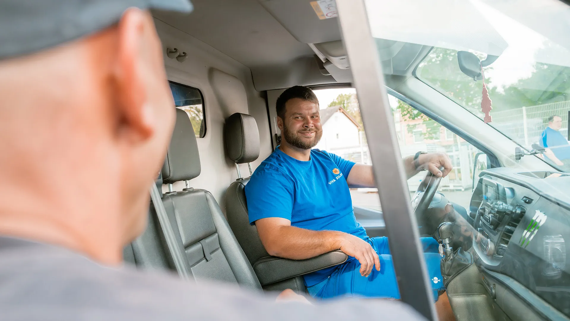 Mann in einem blauen Hemd sitzt in einem Van und lächelt eine andere Person außerhalb an. Die Umgebung ist hell und ungezwungen.