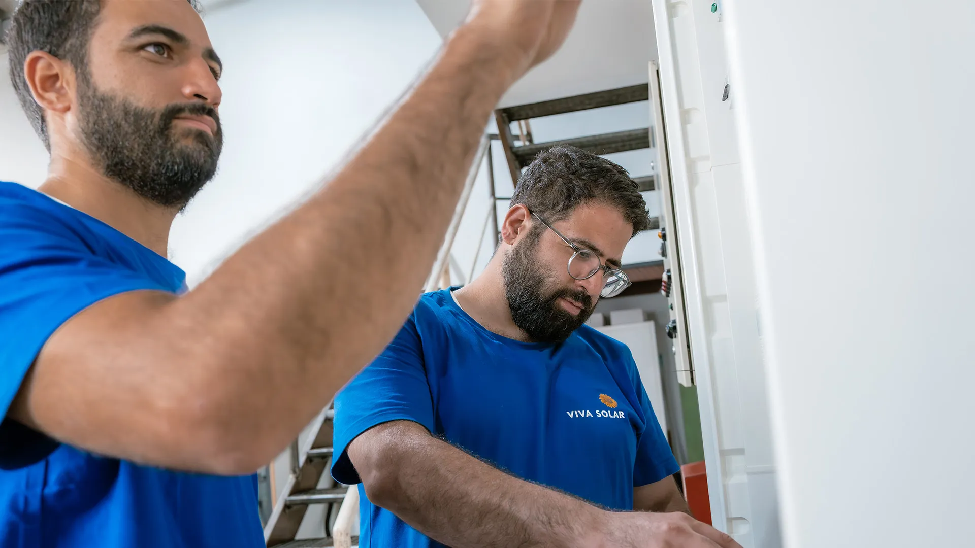 Zwei Männer in blauen Hemden arbeiten in einem Innenraum an Solaranlagen, konzentriert auf ihre Aufgaben, im Hintergrund ist eine Treppe zu sehen.