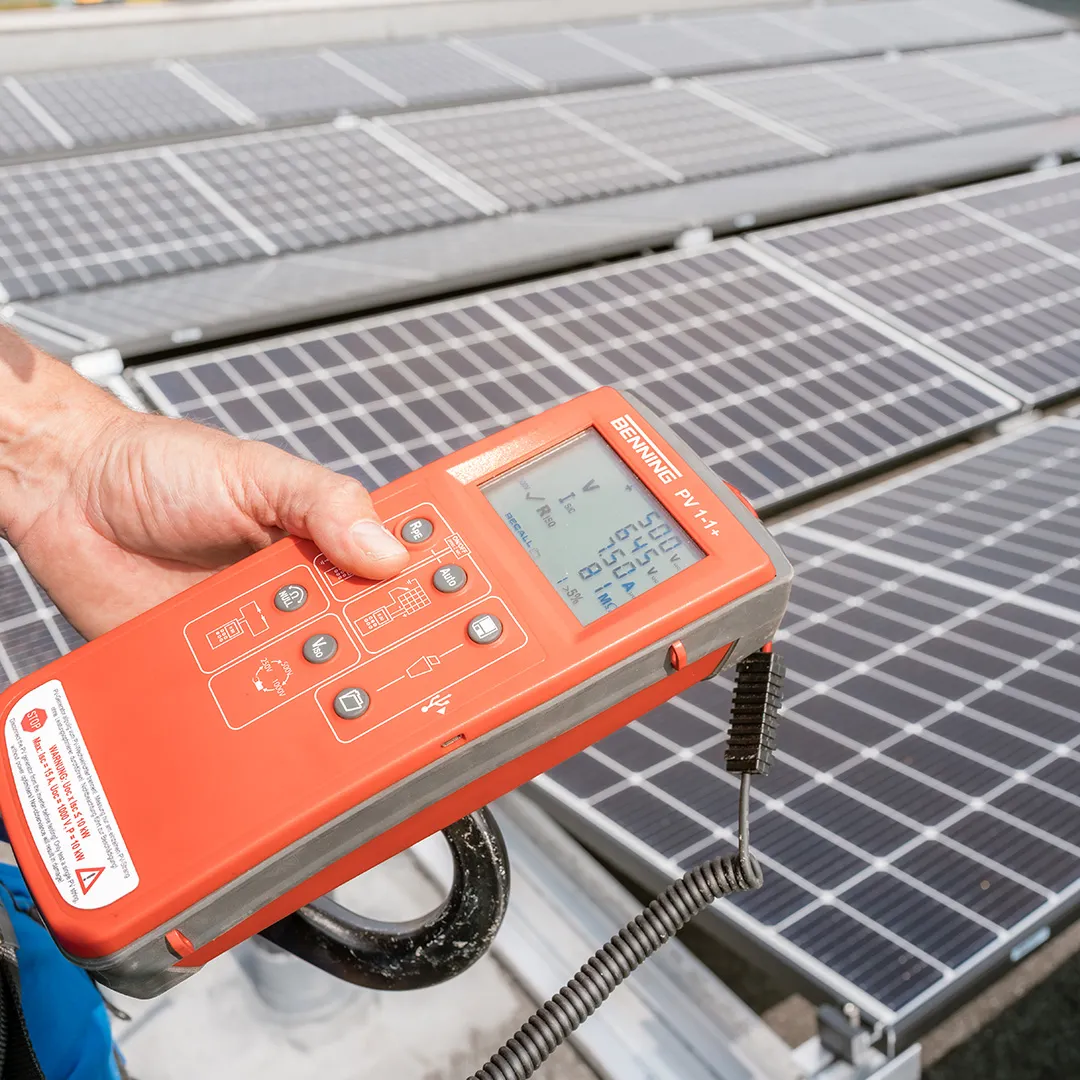 Eine Hand hält einen roten Solarmodul-Tester über Photovoltaikmodule und zeigt Messwerte für Spannung und Strom an.
