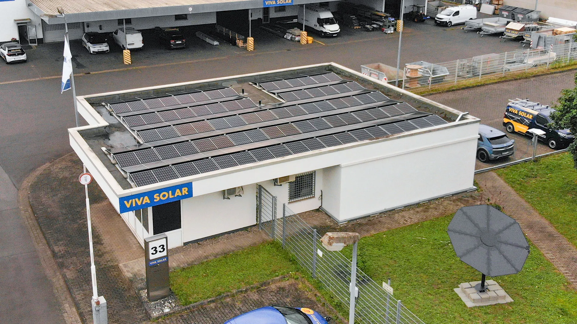Ein kleines weißes Gebäude mit einem blauen Schild mit der Aufschrift „VIVA SOLAR“ hat Sonnenkollektoren auf dem Dach. Umgeben von Grünflächen und geparkten Fahrzeugen.