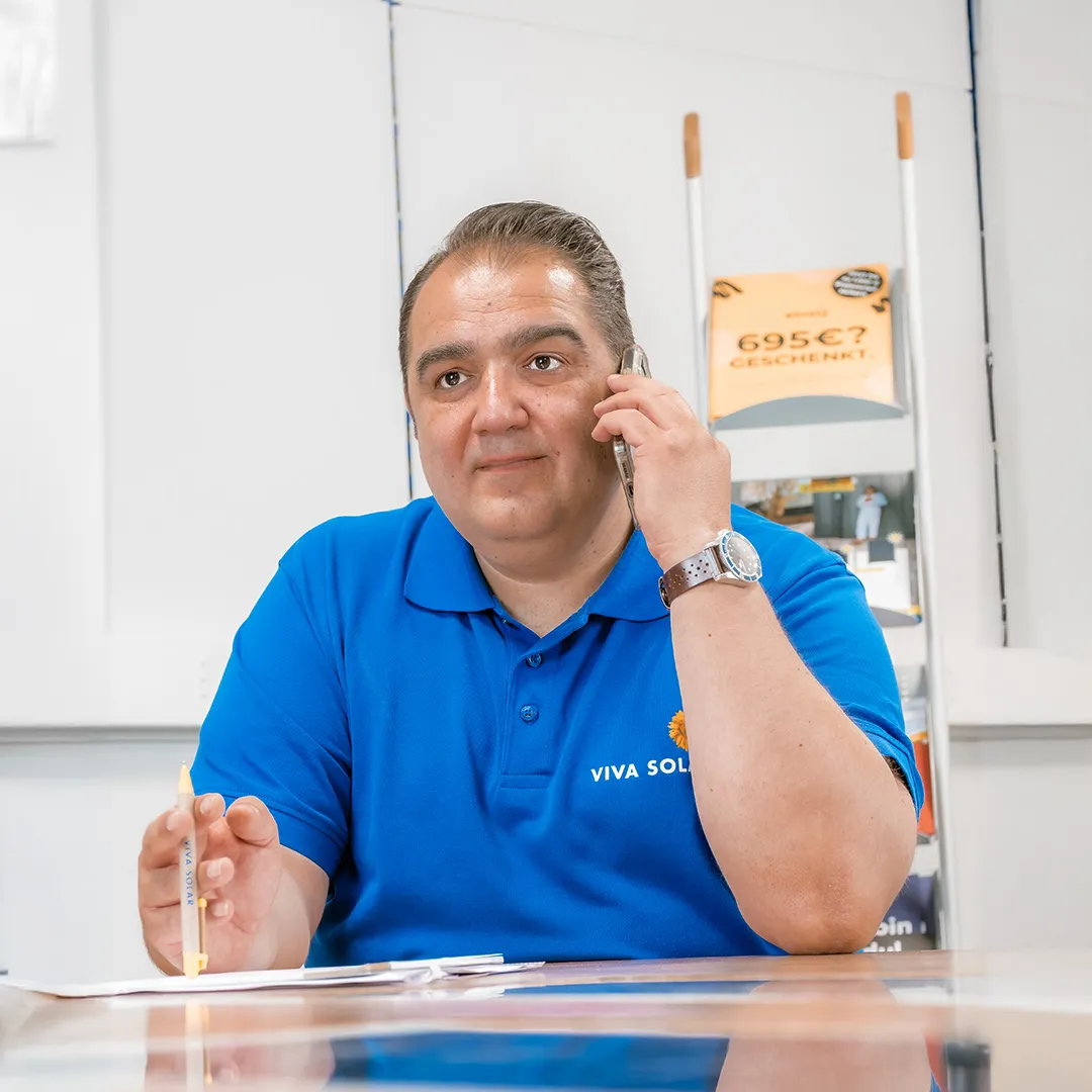 Ein Mann in einem blauen Polohemd telefoniert und hält dabei einen Stift in der Hand. Vor ihm liegen Papiere auf einem Tisch.