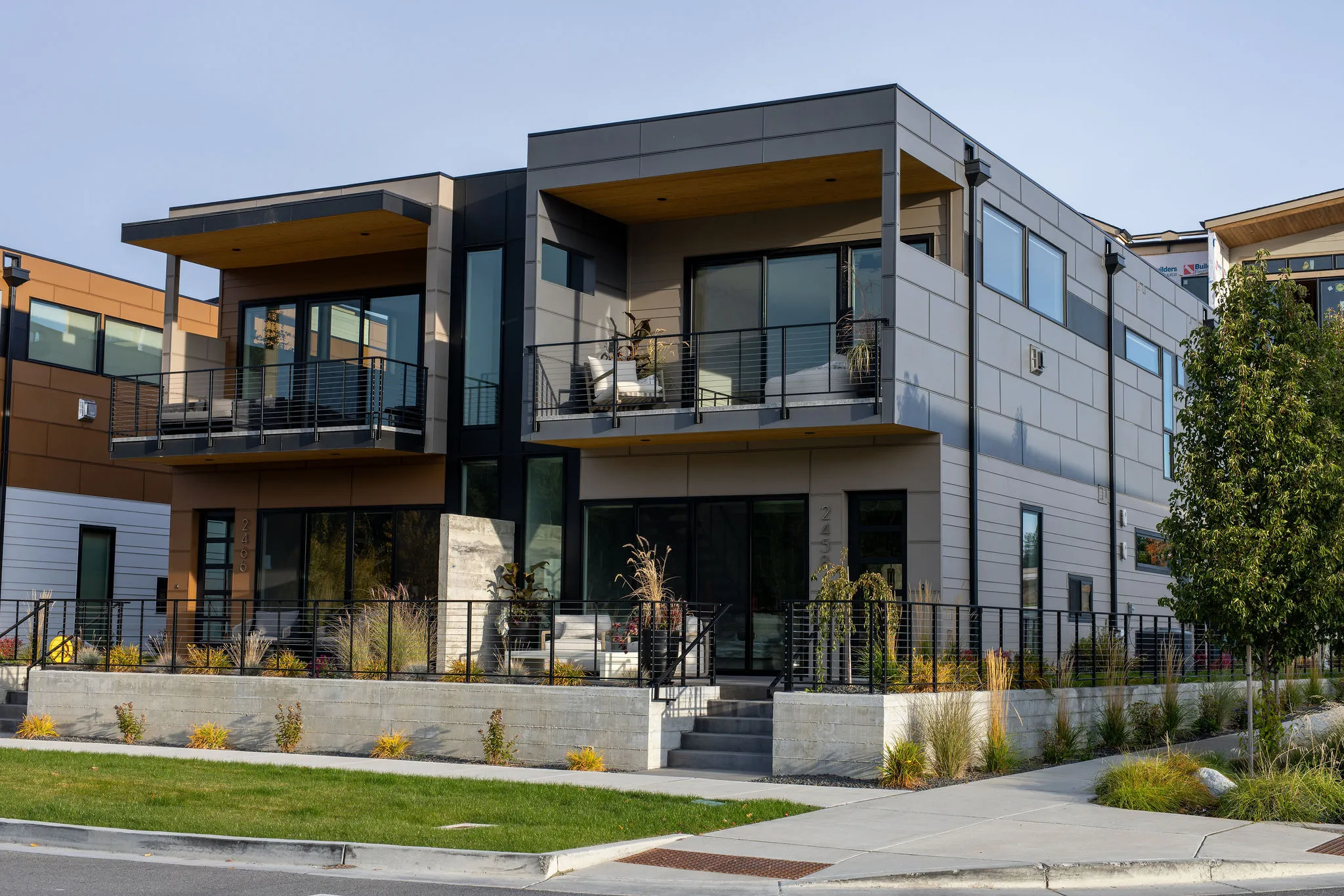 Modern two-story residential building with balconies, large windows, outdoor seating, and landscaped front yard.