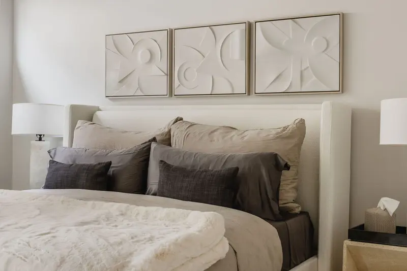 Modern bedroom with a beige upholstered bed, layered gray and beige pillows, a soft beige blanket, abstract wall art above, and two bedside lamps.