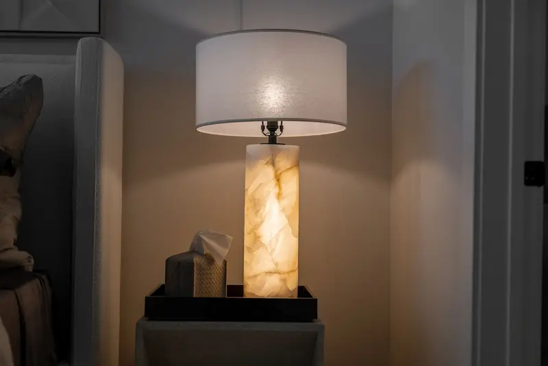 Illuminated table lamp with a marble base and a white shade on a bedside table next to a tissue box.