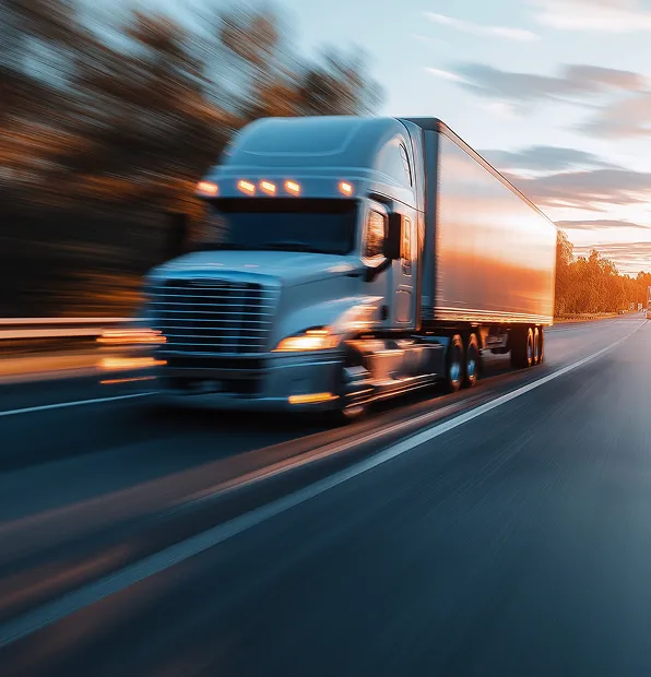 Modern semi-truck with bright front lights speeding down a highway; motion blur emphasizes high velocity, set against a dramatic sunset sky blending orange and deep blue hues.