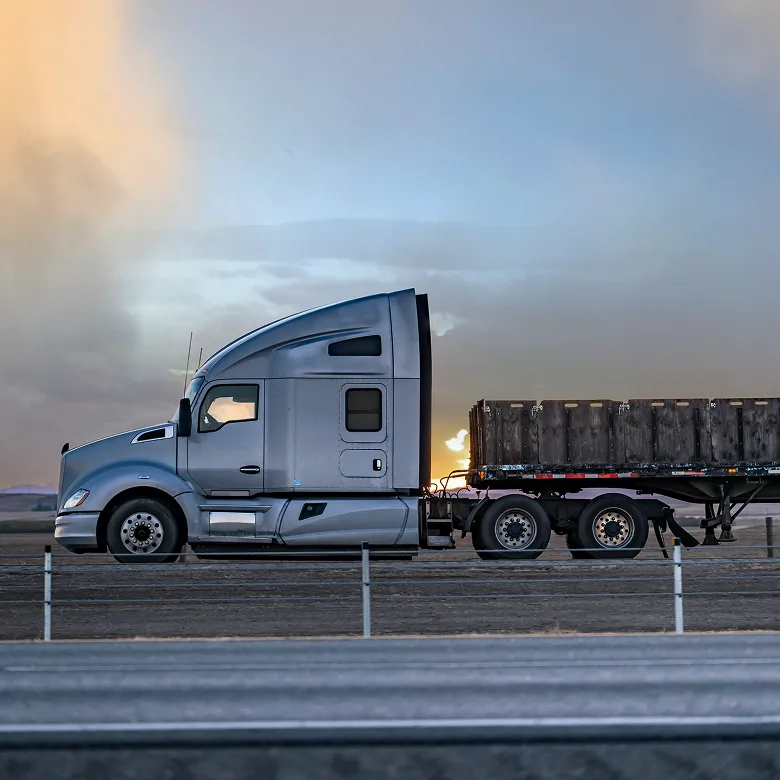 Silver semi-truck with flatbed trailer hauling a large rectangular container, parked on a roadside near a metal guardrail, with a dramatic sky of sunset or sunrise hues in the background.