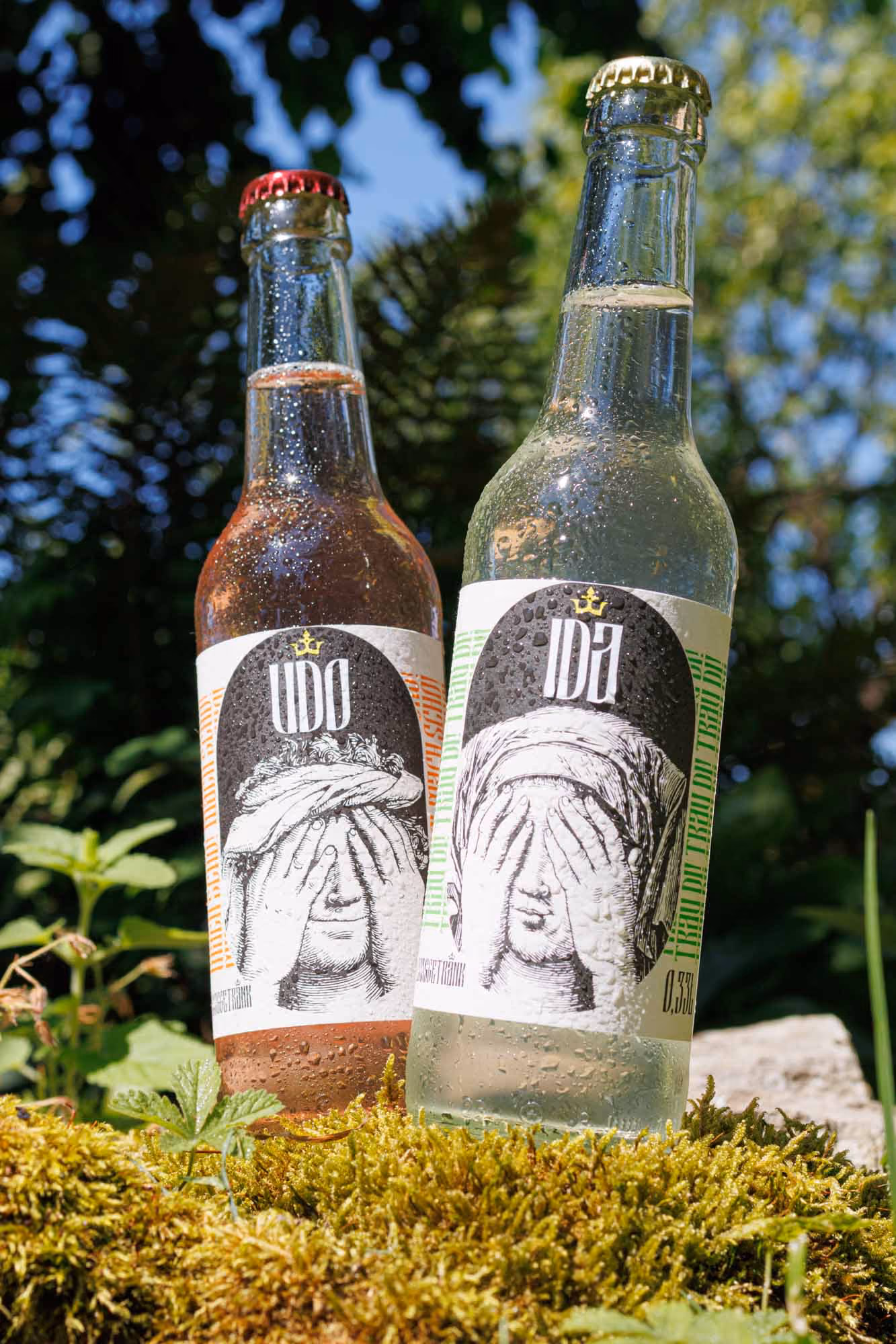 Zwei Flaschen UDO und IDA mit Etiketten, die Figuren zeigen, die sich die Augen mit den Händen bedecken; die Flaschen stehen auf Moos mit Wald und blauem Himmel im Hintergrund.